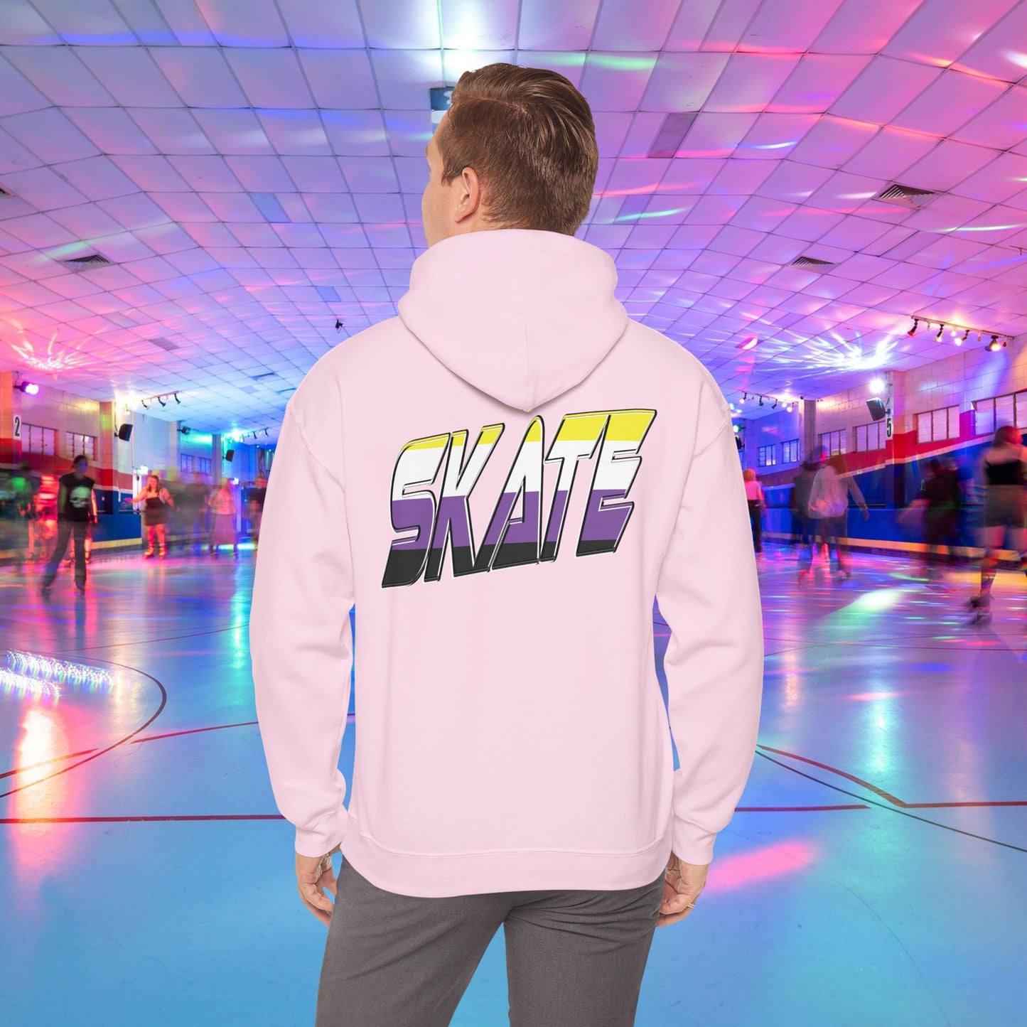 A person with light brown hair is wearing a high-quality SKATE Non-binary Pride Hoodie from Printify, featuring bold, colorful letters spelling "SKATE" on the back in non binary pride flag colours. They are standing in a roller skating rink with flashing neon lights, while other people skate around in the background.