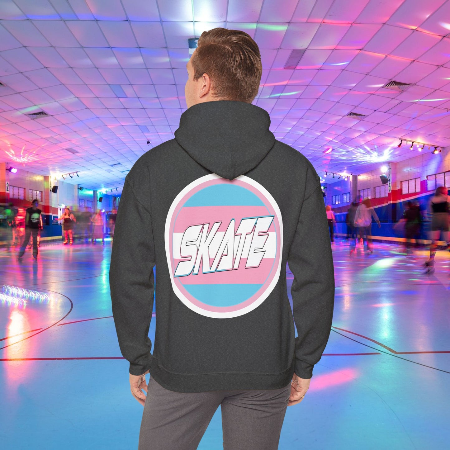 A person wearing a high-quality, unisex "Gay Skate Original - Trans Edition Hoodie" from Printify, featuring a large "SKATE" logo on the back, stands in a colorful roller skating rink. The rink is illuminated with multicolored lights and has several people skating in the background. The overall atmosphere is lively and vibrant.