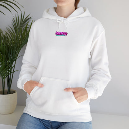 Wearing the loose-fitting Protect Trans Kids Hoodie made from ethically grown cotton, featuring a small, colorful "Protect Your Energy" graphic, a person has hands in the front pocket with a potted plant adding charm to the background.