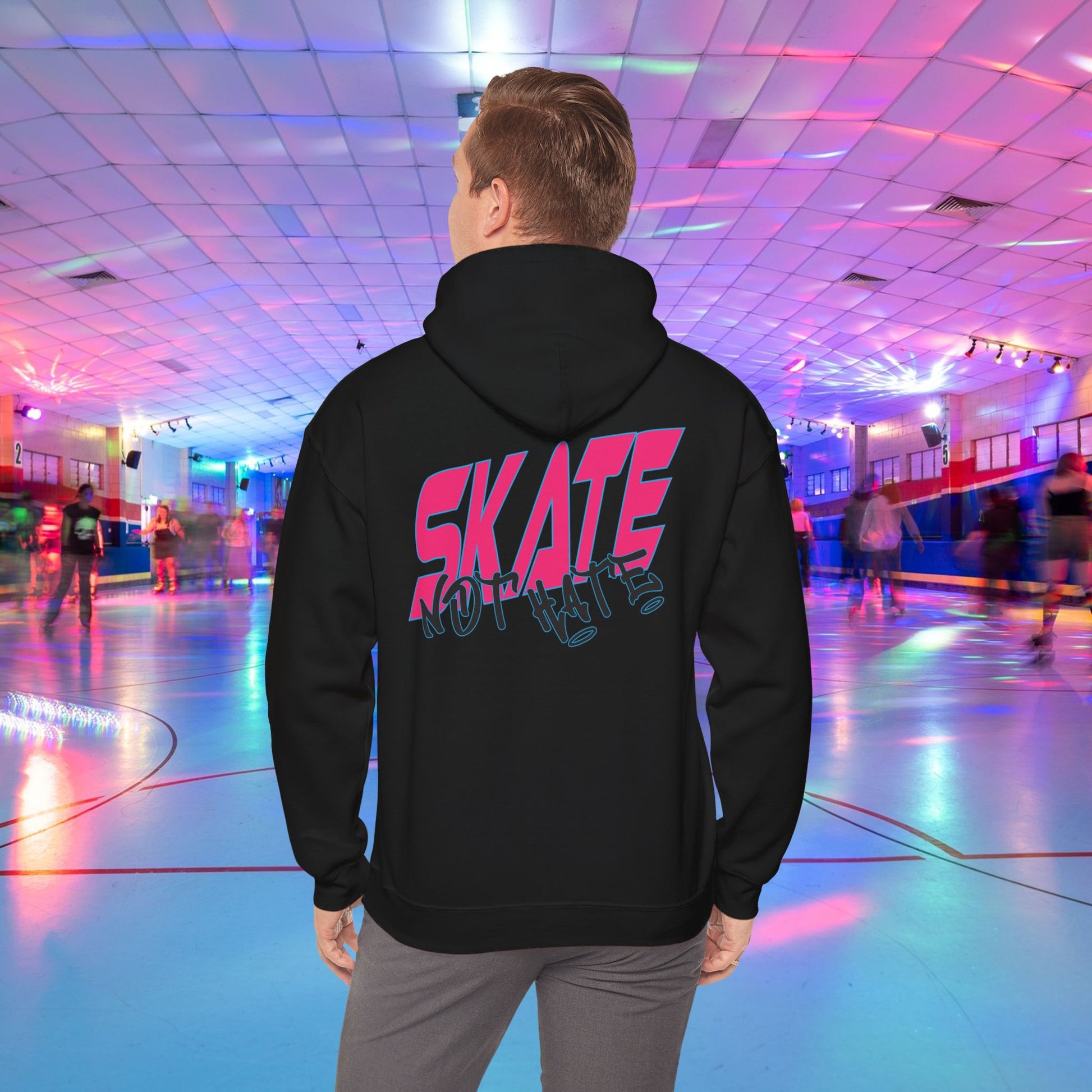 A person wearing a SKATE Not Hate Hoodie from Printify, characterized by its light pink color and high-quality construction, stands in a roller skating rink. The ethically-made unisex heavy blend hoodie, crafted from US-grown cotton, features the words "SKATE NOT HATE" printed on the back. The rink is adorned with colorful lights and has a shiny floor, with other skaters gliding in the background.