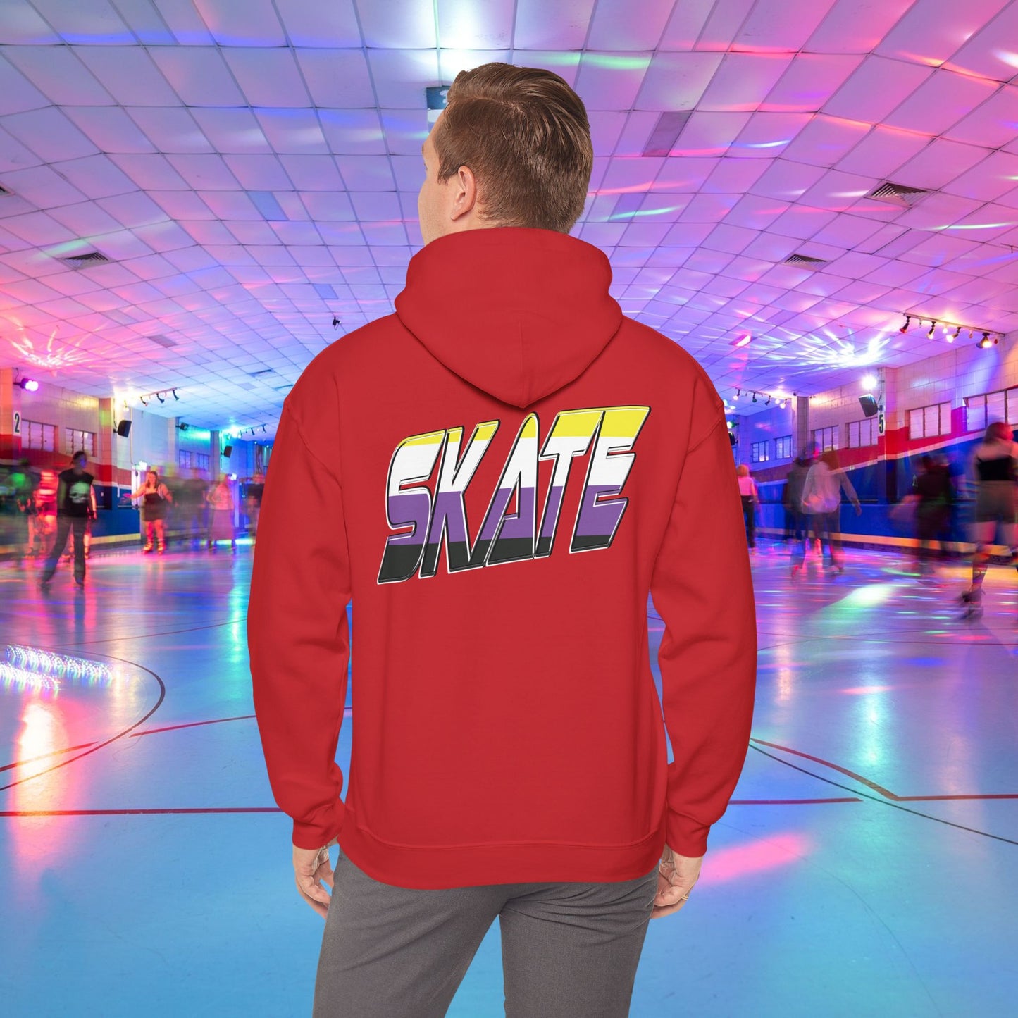 A person with light brown hair is wearing a high-quality SKATE Non-binary Pride Hoodie from Printify, featuring bold, colorful letters spelling "SKATE" on the back in non binary pride flag colours. They are standing in a roller skating rink with flashing neon lights, while other people skate around in the background.