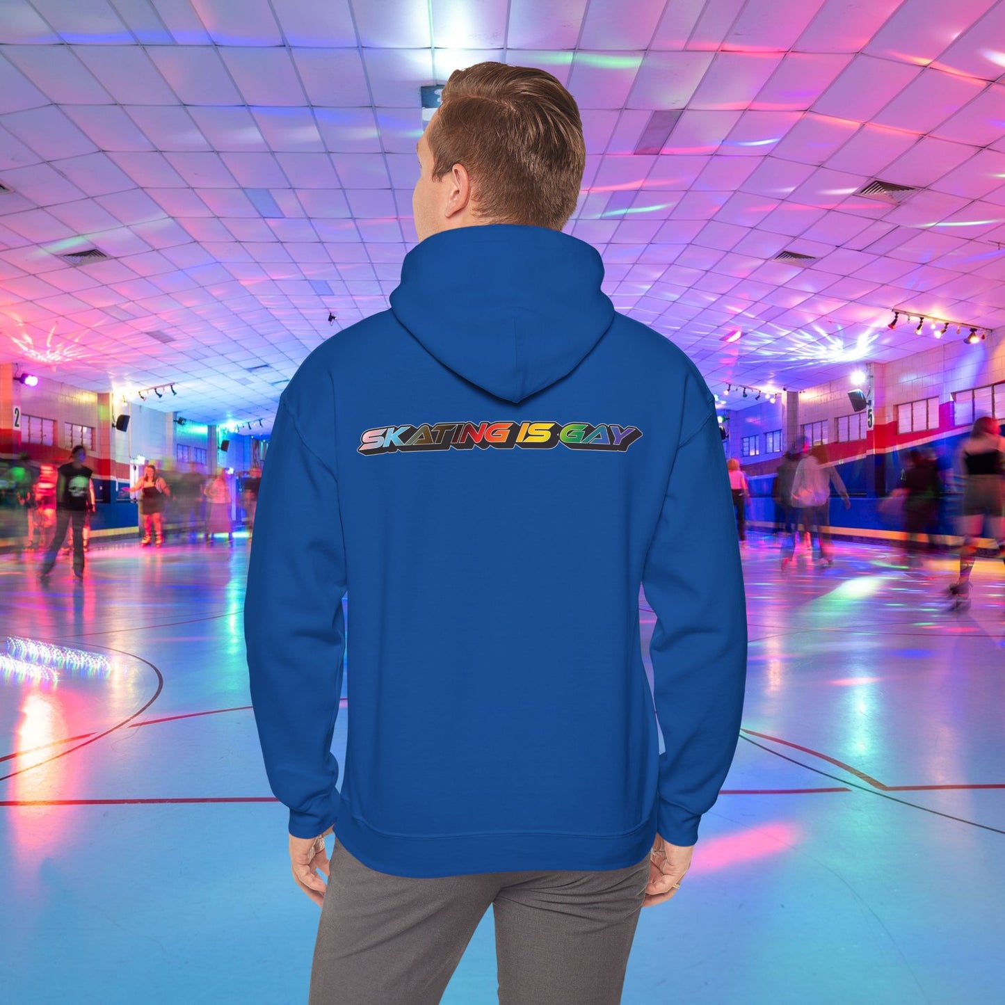 A person wearing a high-quality Skating Is Gay Hoodie by Printify, featuring the vibrant rainbow pride text "SKATING IS GAY" printed on the back, stands in the foreground of a neon-lit roller skating rink. People skate in the blurred background as colorful lights reflect off the rink's surface. The product is available with Australian shipping.