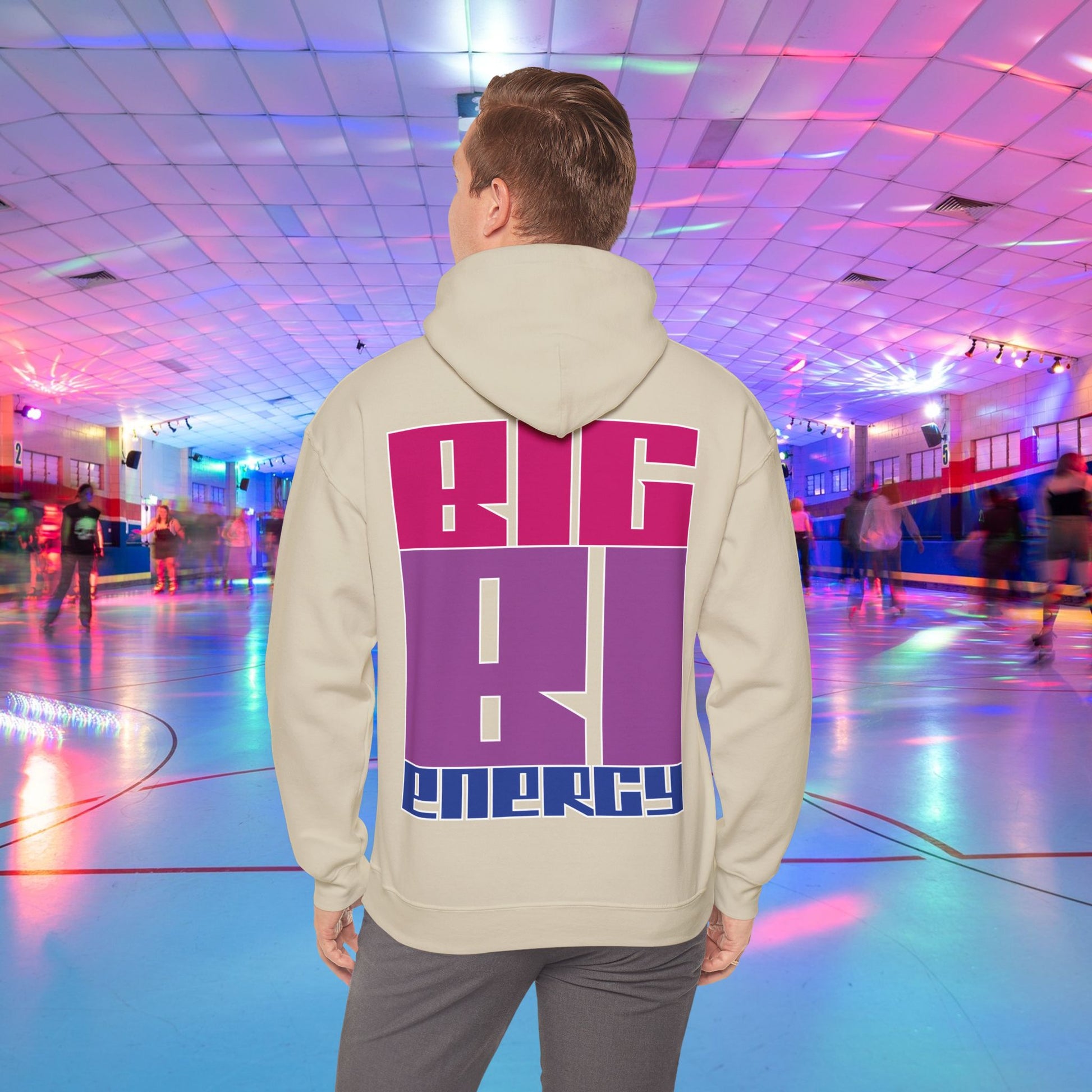 A person with light brown hair is wearing a high-quality BIG BI ENERGY Hoodie from GAY SKATE, featuring bold, Bisexual Pride Flag colour letters on the back. They are standing in a roller skating rink illuminated with vibrant, colorful lights. The cozy feel and warmth of this trans and queer-owned brand is evident.