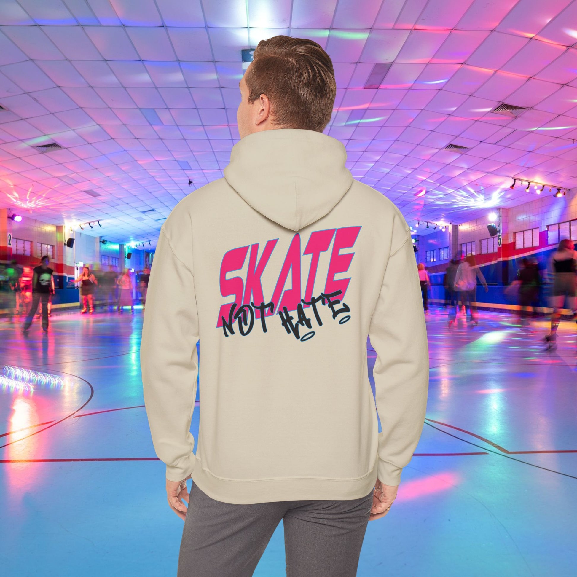 A person wearing a SKATE Not Hate Hoodie from Printify, characterized by its light pink color and high-quality construction, stands in a roller skating rink. The ethically-made unisex heavy blend hoodie, crafted from US-grown cotton, features the words "SKATE NOT HATE" printed on the back. The rink is adorned with colorful lights and has a shiny floor, with other skaters gliding in the background.