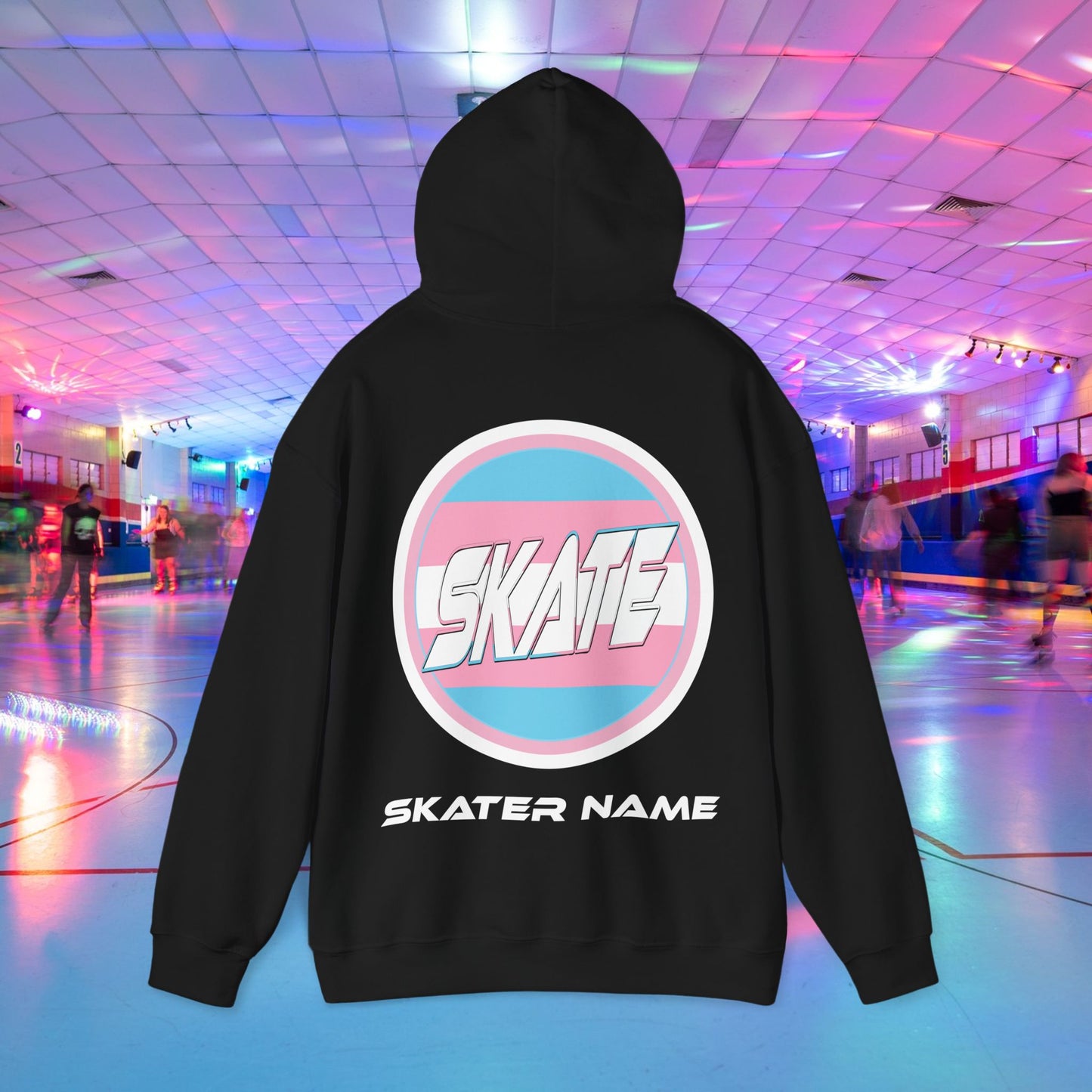 A person wearing a Printify "Add Custom Name - SKATE Trans Flag round logo Hoodie - Australian Shipping," a black unisex heavy blend hooded sweatshirt featuring the word "SKATE" inside a trans circle logo on the back with their skater name written beneath it. The background shows an indoor roller skating rink with colorful lights and other skaters.