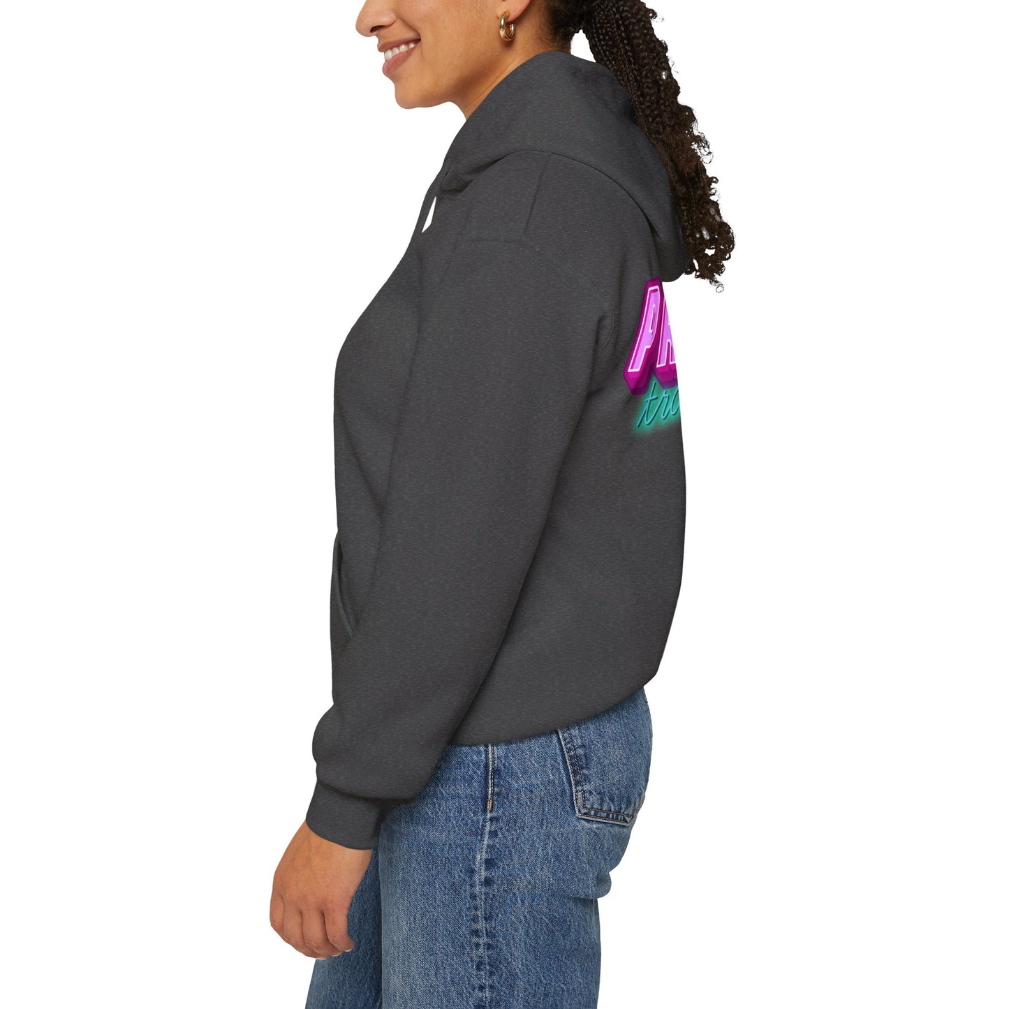 Wearing the unisex Protect Trans Kids Hoodie in dark gray, featuring colorful back text, a person stands in profile. Their curly hair is pulled back, and they wear it with blue jeans. Crafted from ethically grown cotton, the hoodie contrasts against the plain white background.