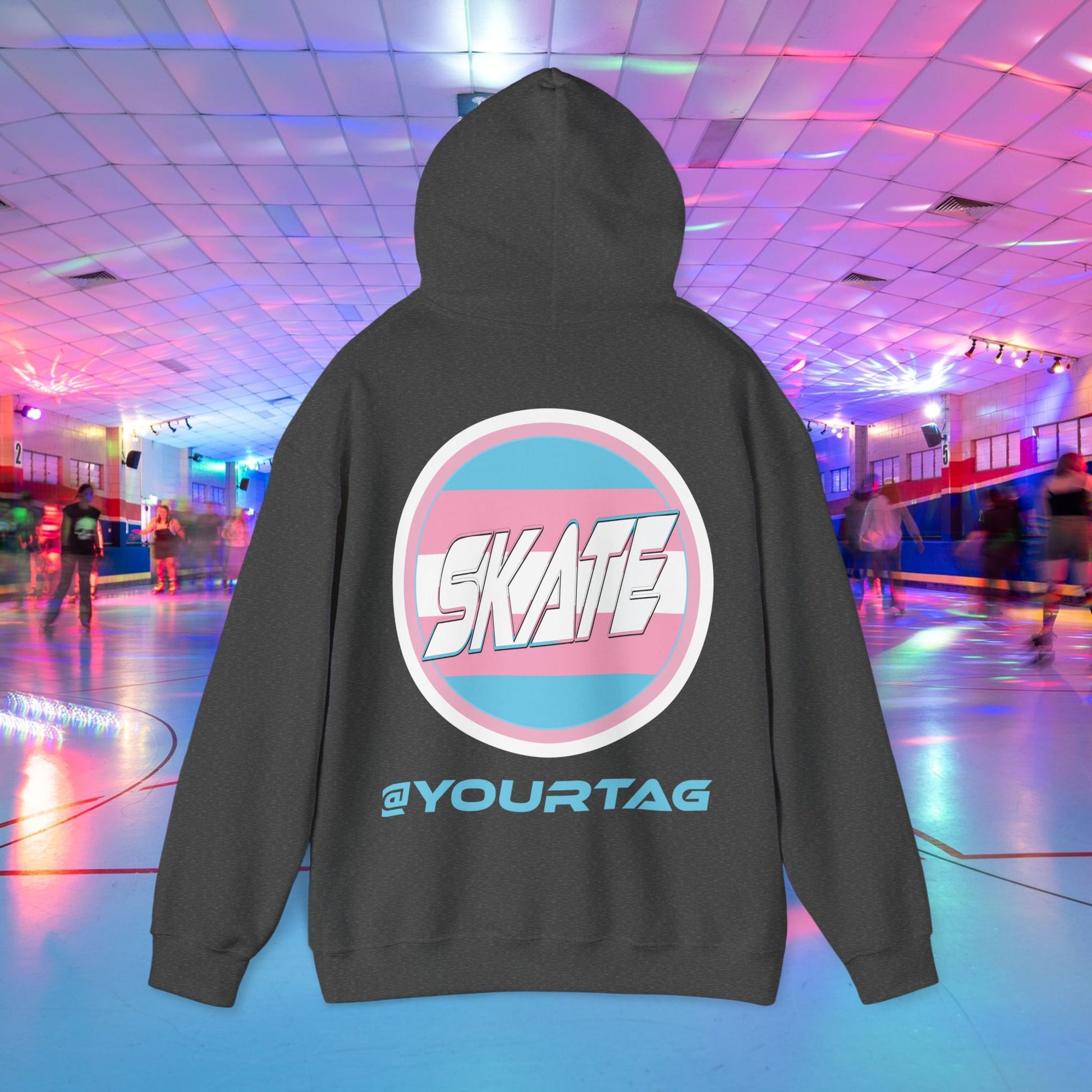 A person wearing Printify's "Add Custom Name - Trans Edition Hoodie" is showcasing their unique style at a roller skating rink. The dark hoodie features a large "SKATE" logo and the customizable text "@YOURTAG" on the back, making them stand out amidst the neon lights and bustling crowd of skaters.