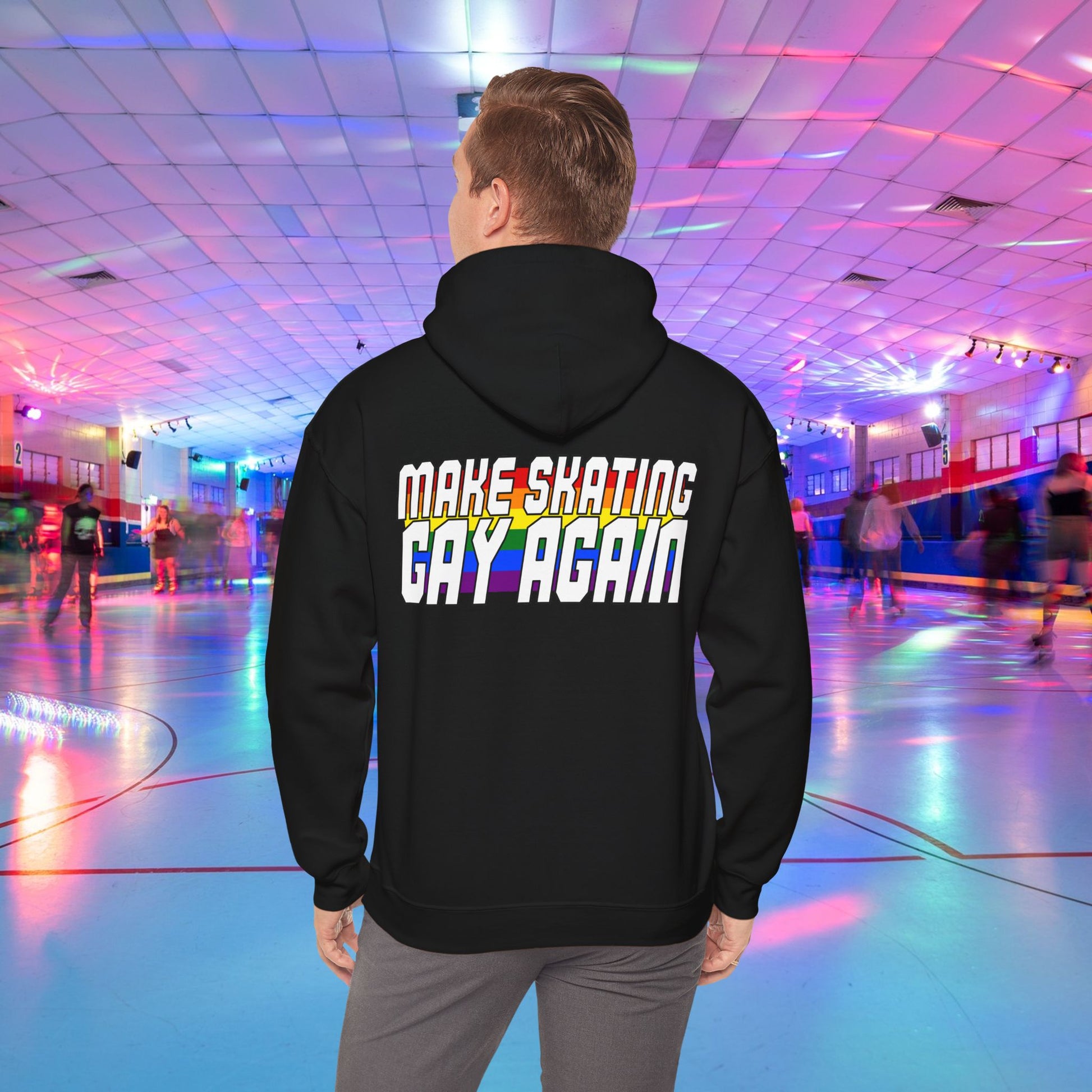 A person, seen from behind, is wearing the "Make Skating Gay Again Rainbow Roller Skate Love Heart Hoodie" from Gay Skate. The hoodie features the phrase "MAKE SKATING GAY AGAIN" in colorful, bold letters. They are in a roller skating rink with vibrant lights. The front features a rainbow pride love heart with a roller skate shaped cut out.
