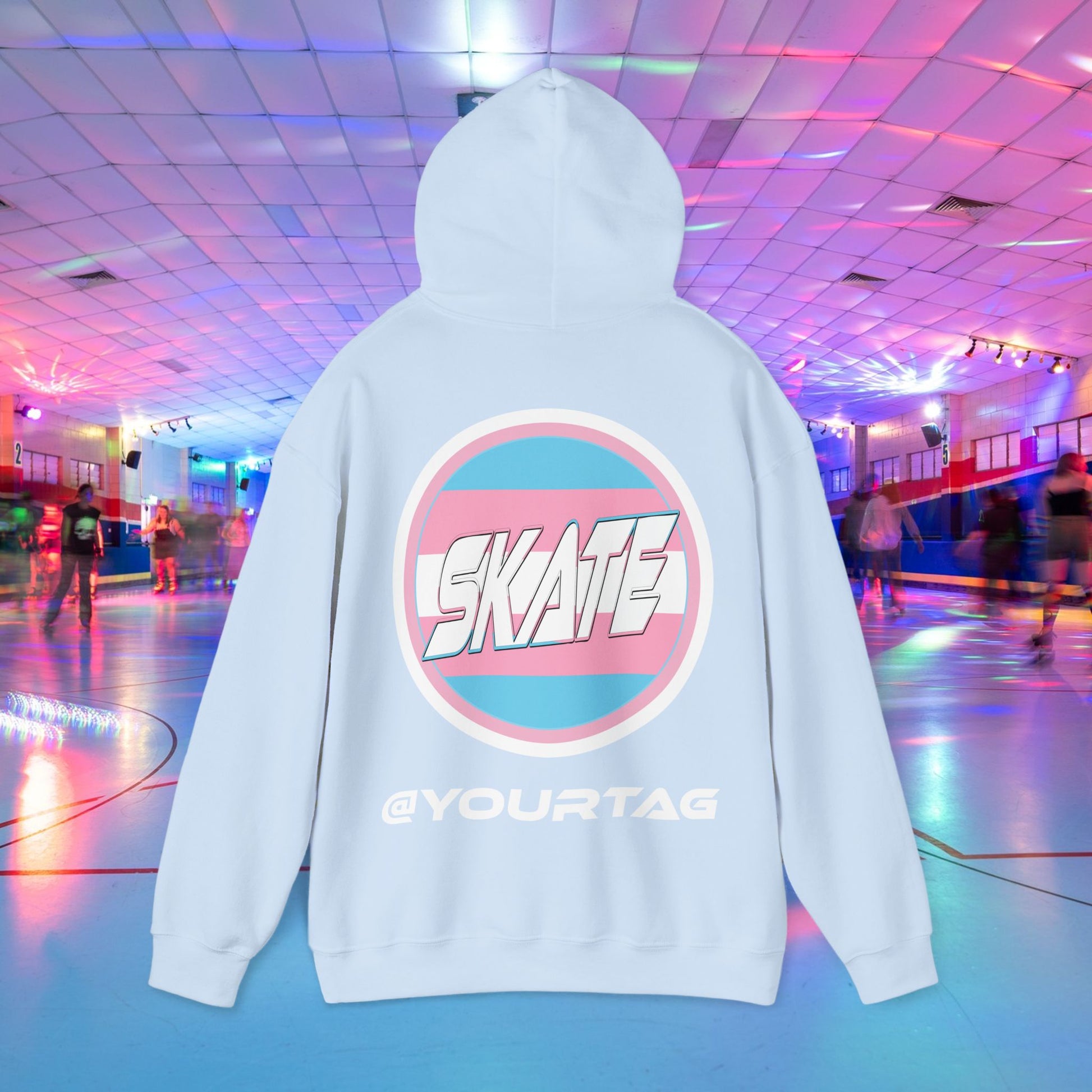 A person wearing a Printify "Add Custom Name - SKATE Trans Flag round logo Hoodie - Australian Shipping," a black unisex heavy blend hooded sweatshirt featuring the word "SKATE" inside a trans circle logo on the back with their skater name written beneath it. The background shows an indoor roller skating rink with colorful lights and other skaters.