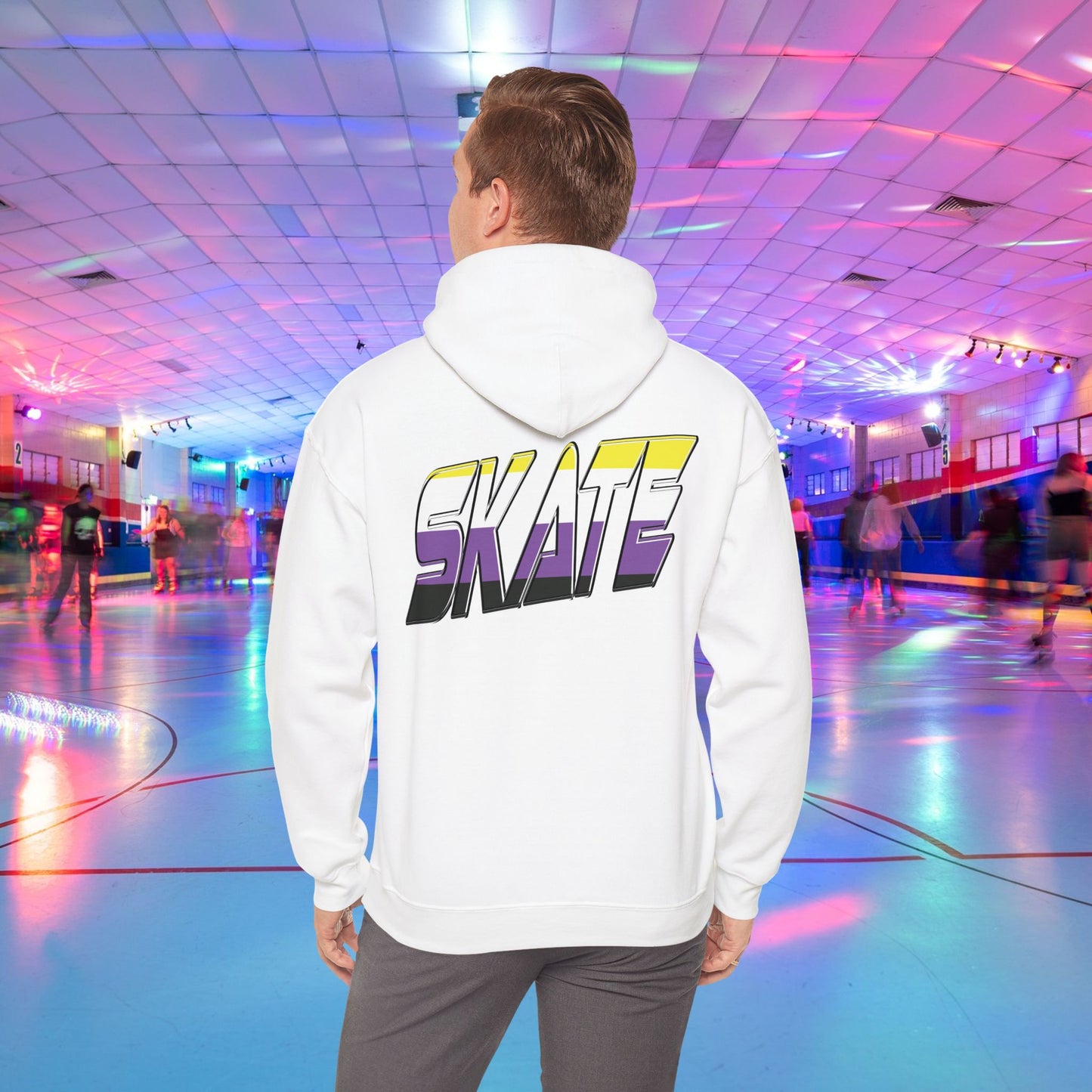 A person with light brown hair is wearing a high-quality SKATE Non-binary Pride Hoodie from Printify, featuring bold, colorful letters spelling "SKATE" on the back in non binary pride flag colours. They are standing in a roller skating rink with flashing neon lights, while other people skate around in the background.