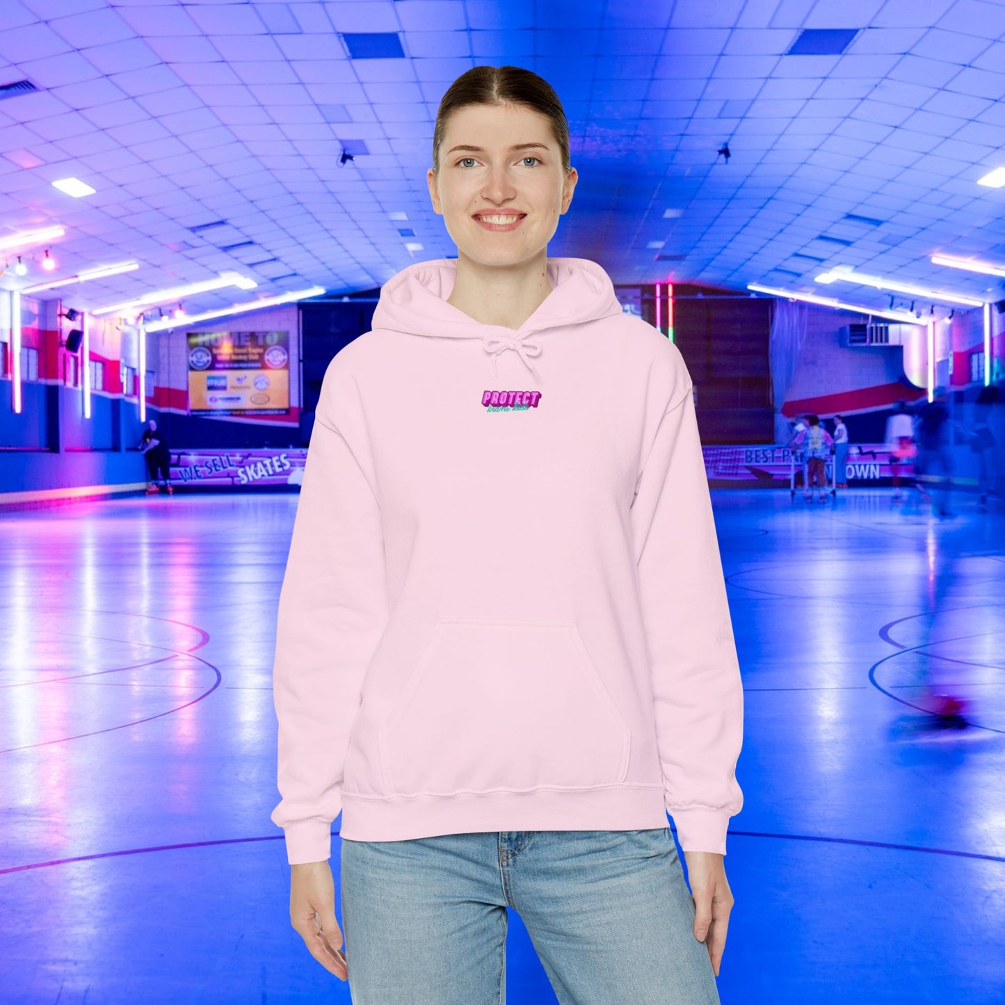 Wearing a "Protect Trans Kids" hoodie made from ethically grown cotton, a person smiles while standing in an indoor roller skating rink. Blue and pink lights illuminate the background with blurry figures skating.