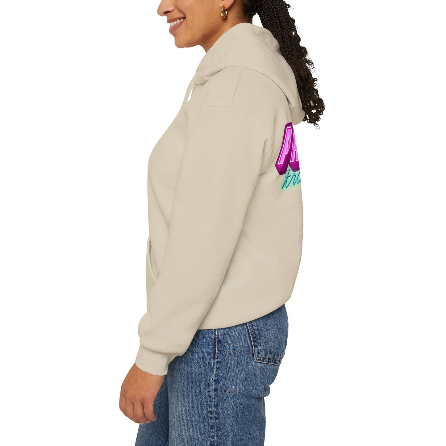 A person with long curly hair wears the beige "Protect Trans Kids Hoodie - Australian Shipping" featuring colorful lettering on the back and blue jeans. They smile, standing against a plain white background, shown from the side.