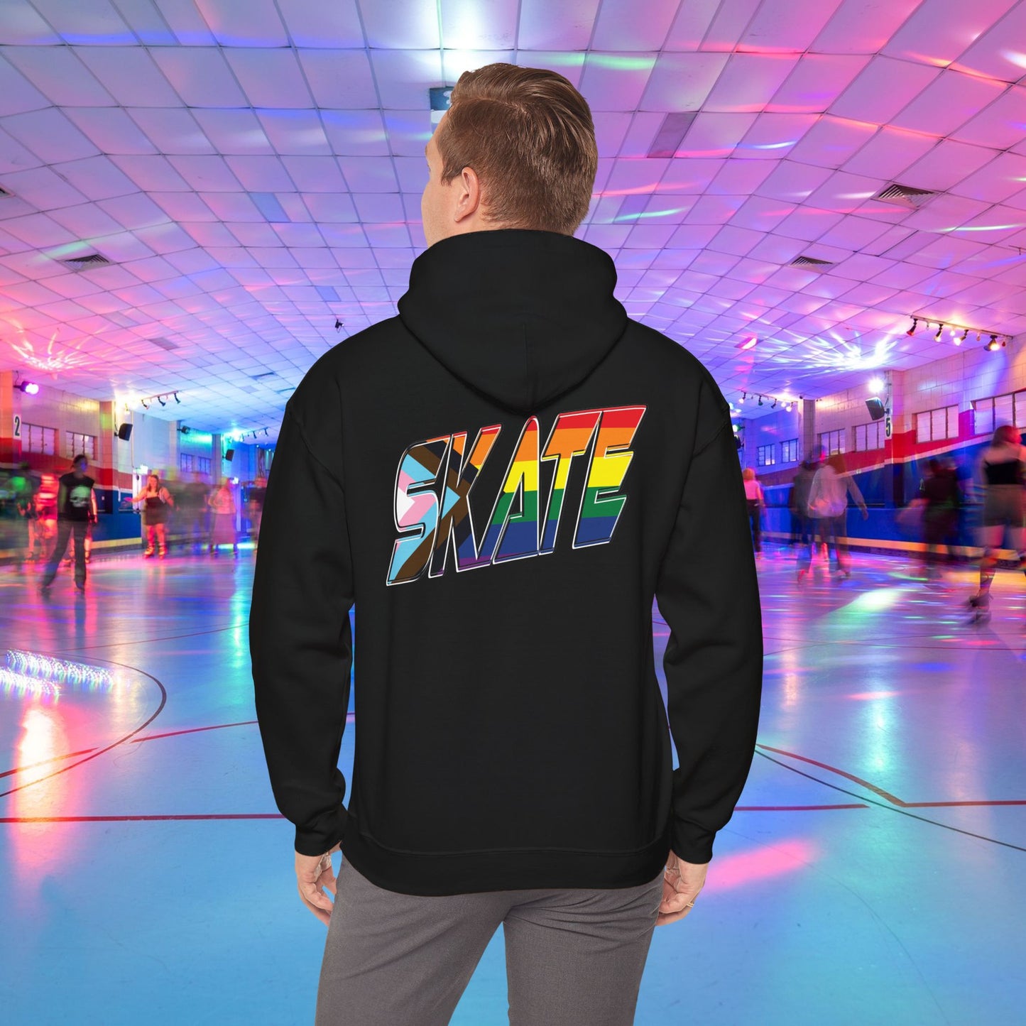 A person with short hair is wearing a GAYSKATE SKATE Progress Pride Hoodie, showcasing a rainbow pride flag gradient "SKATE" design on the back. They are facing away from the camera in a roller skating rink illuminated by vibrant, colorful lights and surrounded by other skaters.