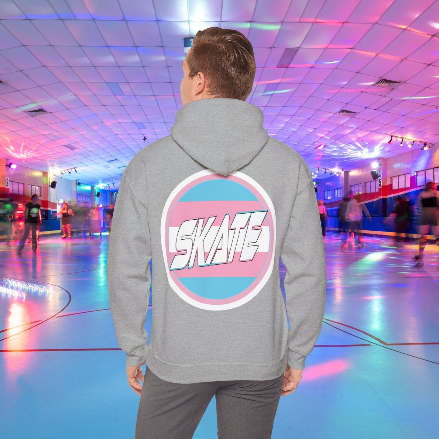 A person wearing a high-quality, unisex "Gay Skate Original - Trans Edition Hoodie" from Printify, featuring a large "SKATE" logo on the back, stands in a colorful roller skating rink. The rink is illuminated with multicolored lights and has several people skating in the background. The overall atmosphere is lively and vibrant.