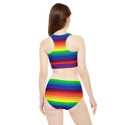 A two-piece "Sporty Swim or Activewear Set - Rainbow Pride Flag" from Printify lays flat against a white background. It features horizontal rainbow stripes in vibrant colors such as red, orange, yellow, green, blue, and purple. The top boasts a high neckline while the bottom has a flattering high-waisted cut.