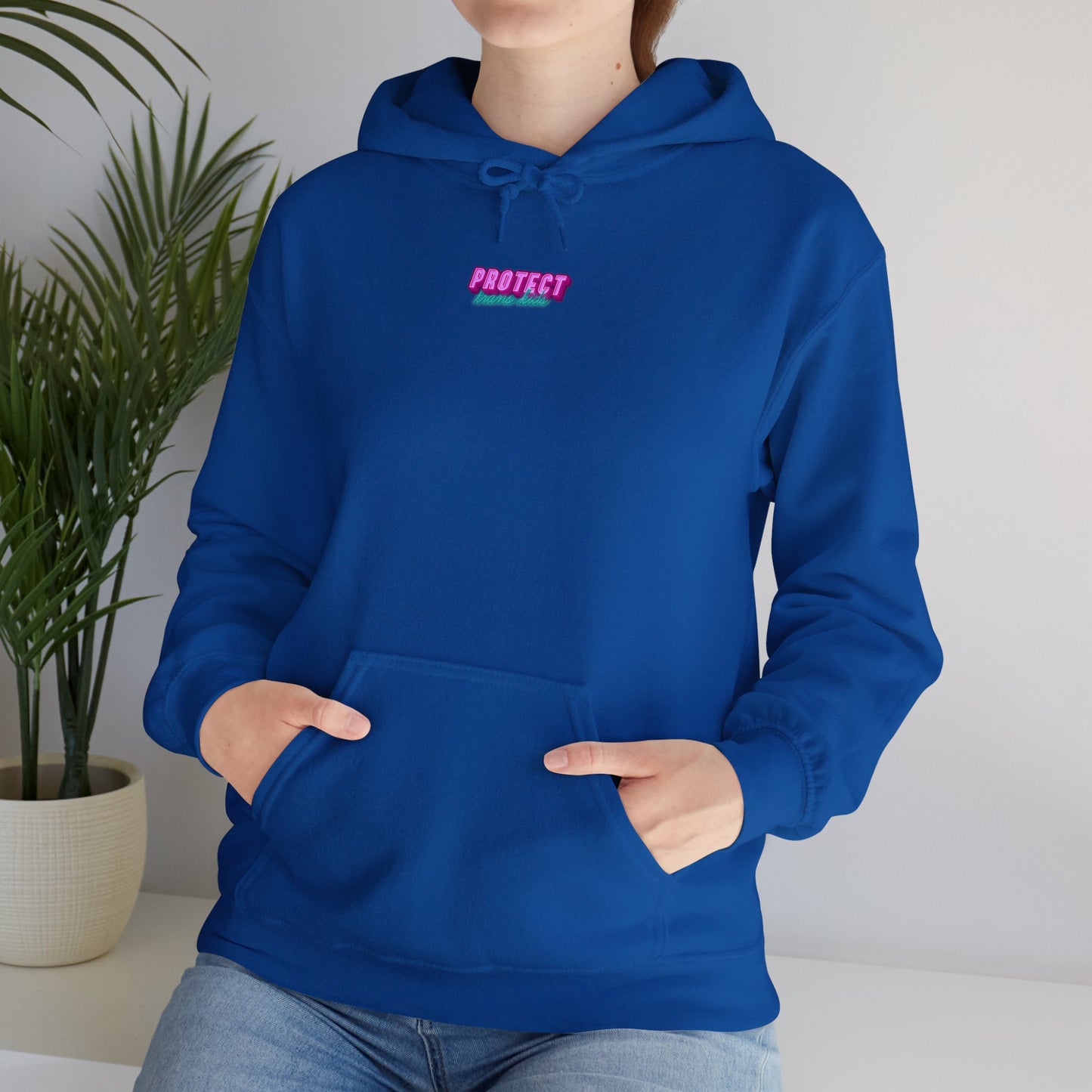 A person wears a blue "Protect Trans Kids" unisex heavy blend sweatshirt featuring "Protect" in pink and a front pocket, with hands nestled inside. A potted plant enhances the natural backdrop.