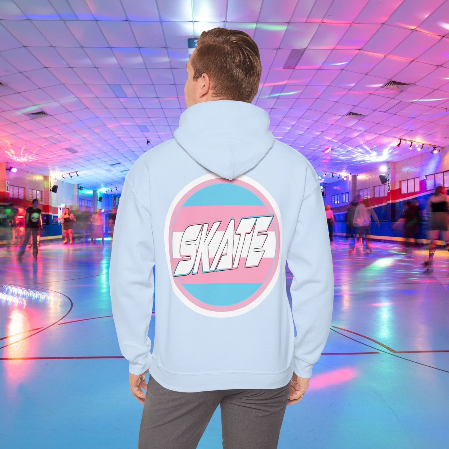 A person wearing a Printify SKATE Trans Flag round logo Hoodie, featuring the word "SKATE" inside a blue and pink circular graphic, stands at the center of a colorful roller skating rink with vibrant lights and other skaters in the background.