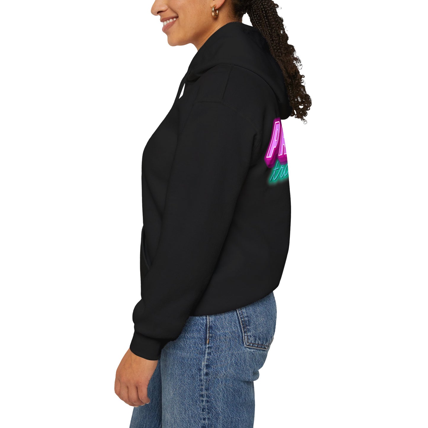 A person is smiling, turned slightly sideways, and wearing a Protect Trans Kids Hoodie - Australian Shipping in black with colorful text on the back, paired with blue jeans against a plain white background.