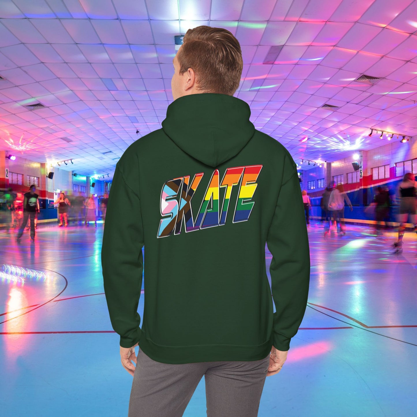 A person with short hair is wearing a GAYSKATE SKATE Progress Pride Hoodie, showcasing a rainbow pride flag gradient "SKATE" design on the back. They are facing away from the camera in a roller skating rink illuminated by vibrant, colorful lights and surrounded by other skaters.