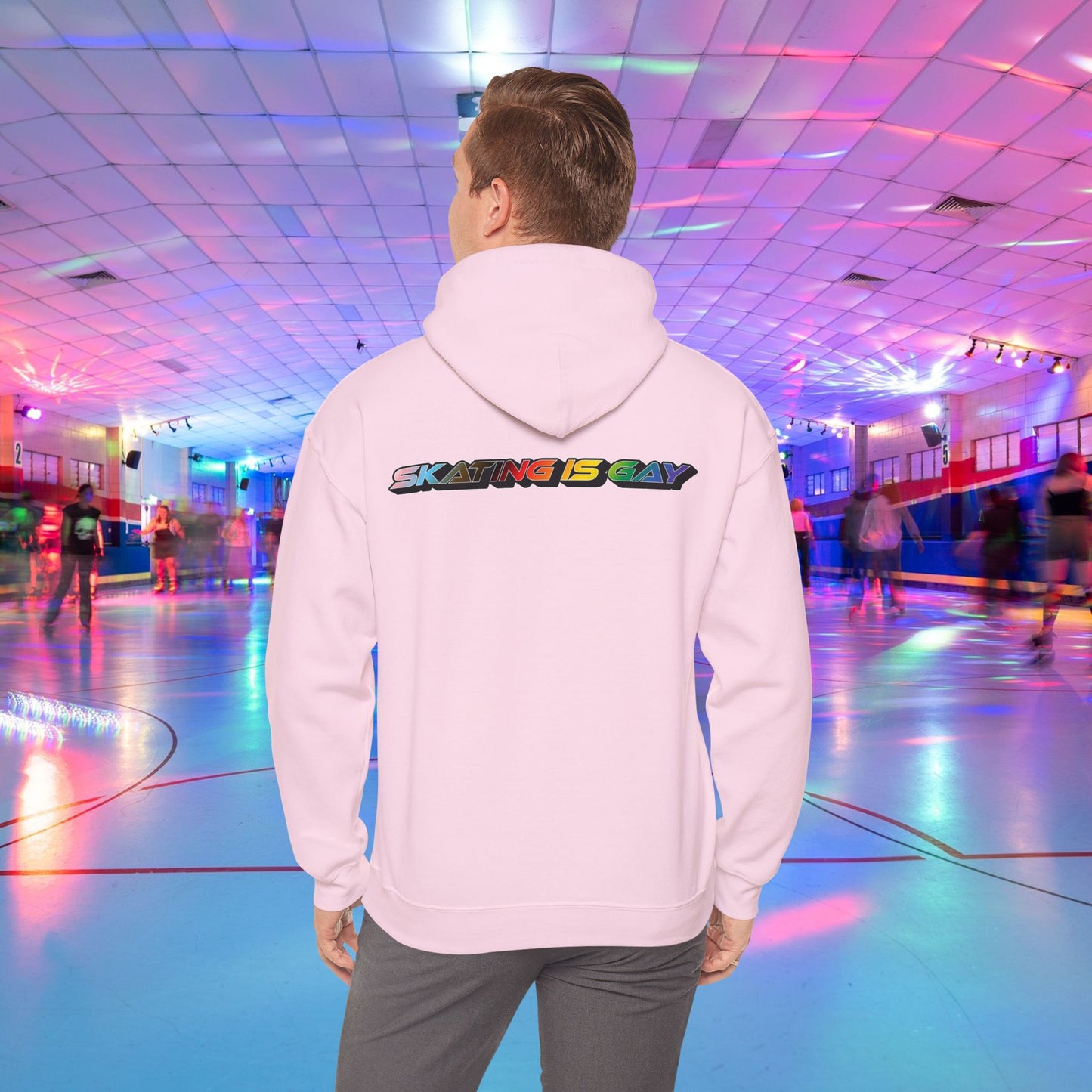 A person wearing a high-quality Skating Is Gay Hoodie by Printify, featuring the vibrant rainbow pride text "SKATING IS GAY" printed on the back, stands in the foreground of a neon-lit roller skating rink. People skate in the blurred background as colorful lights reflect off the rink's surface. The product is available with Australian shipping.