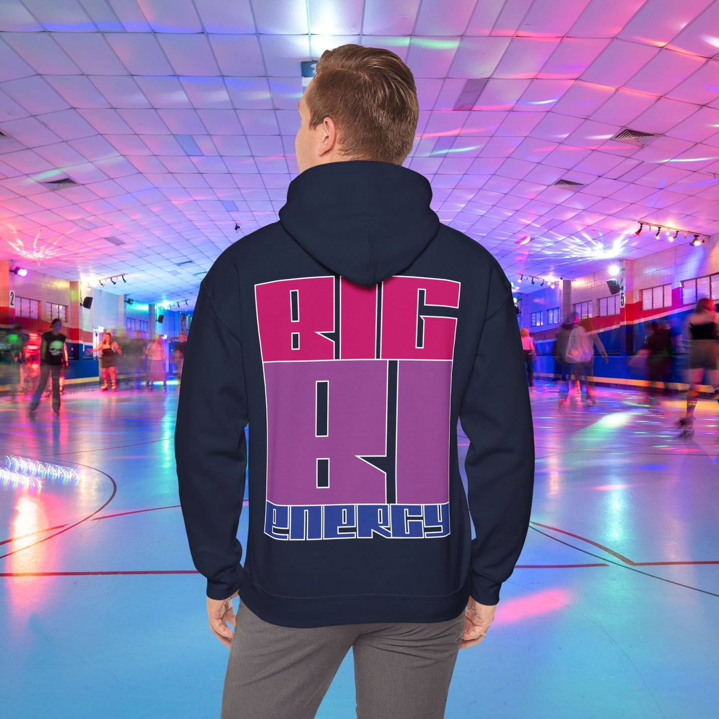 A person with light brown hair is wearing a high-quality BIG BI ENERGY Hoodie from GAY SKATE, featuring bold, Bisexual Pride Flag colour letters on the back. They are standing in a roller skating rink illuminated with vibrant, colorful lights. The cozy feel and warmth of this trans and queer-owned brand is evident.