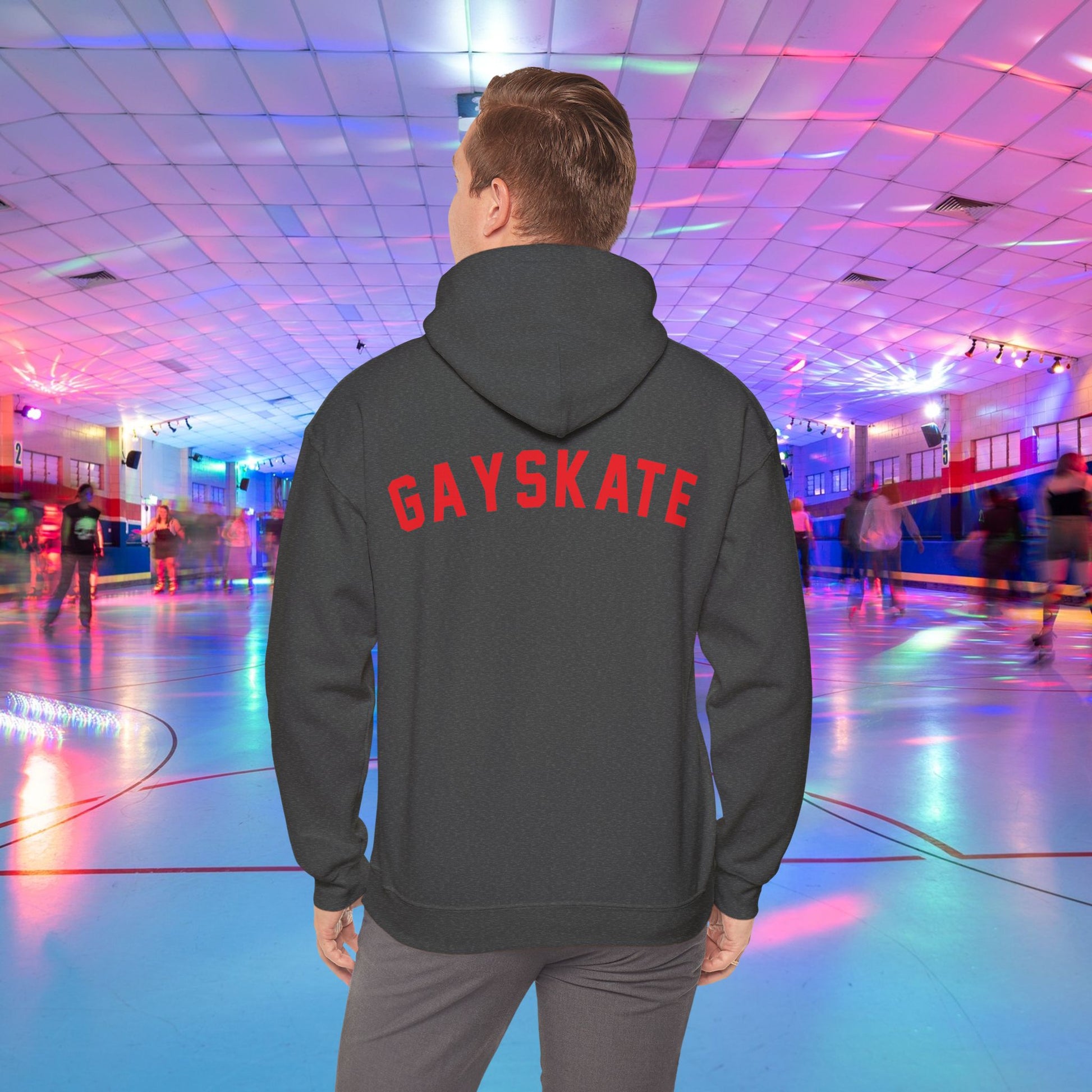 A person wearing a Printify Gay Skate 1982 Retro Hoodie, a high-quality unisex heavy blend hooded sweatshirt, with the word "GAYSKATE" in bold red letters on the back stands in a brightly lit roller skating rink. The rink features colorful lights and skaters in motion, creating a lively atmosphere.