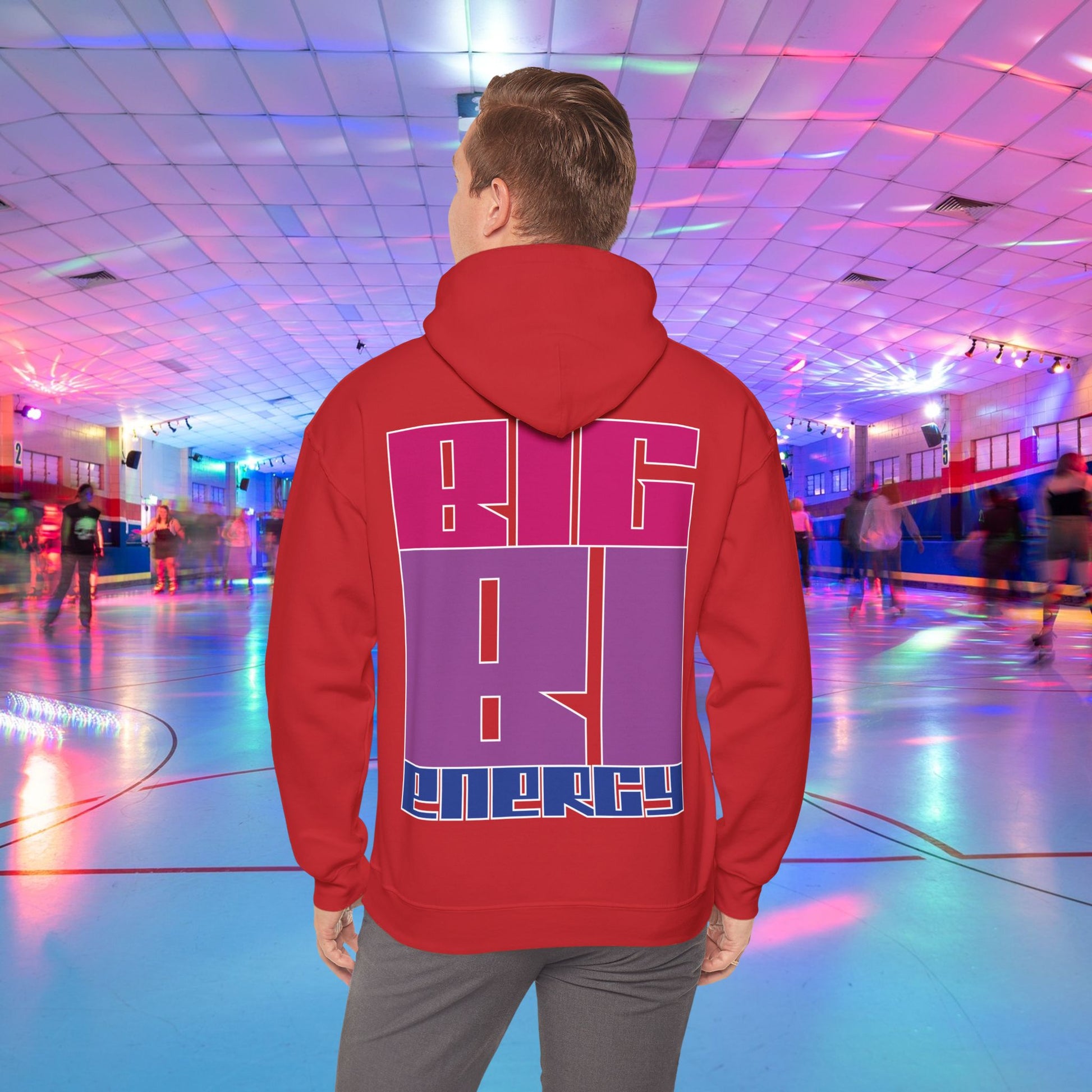 A person with light brown hair is wearing a high-quality BIG BI ENERGY Hoodie from GAY SKATE, featuring bold, Bisexual Pride Flag colour letters on the back. They are standing in a roller skating rink illuminated with vibrant, colorful lights. The cozy feel and warmth of this trans and queer-owned brand is evident.