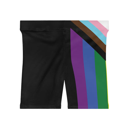 A person is wearing Progress Pride Bike Shorts by Printify, which feature a colorful design with a rainbow pride flag incorporating stripes for people of color and the transgender community. These moisture-wicking shorts fit closely to the body.