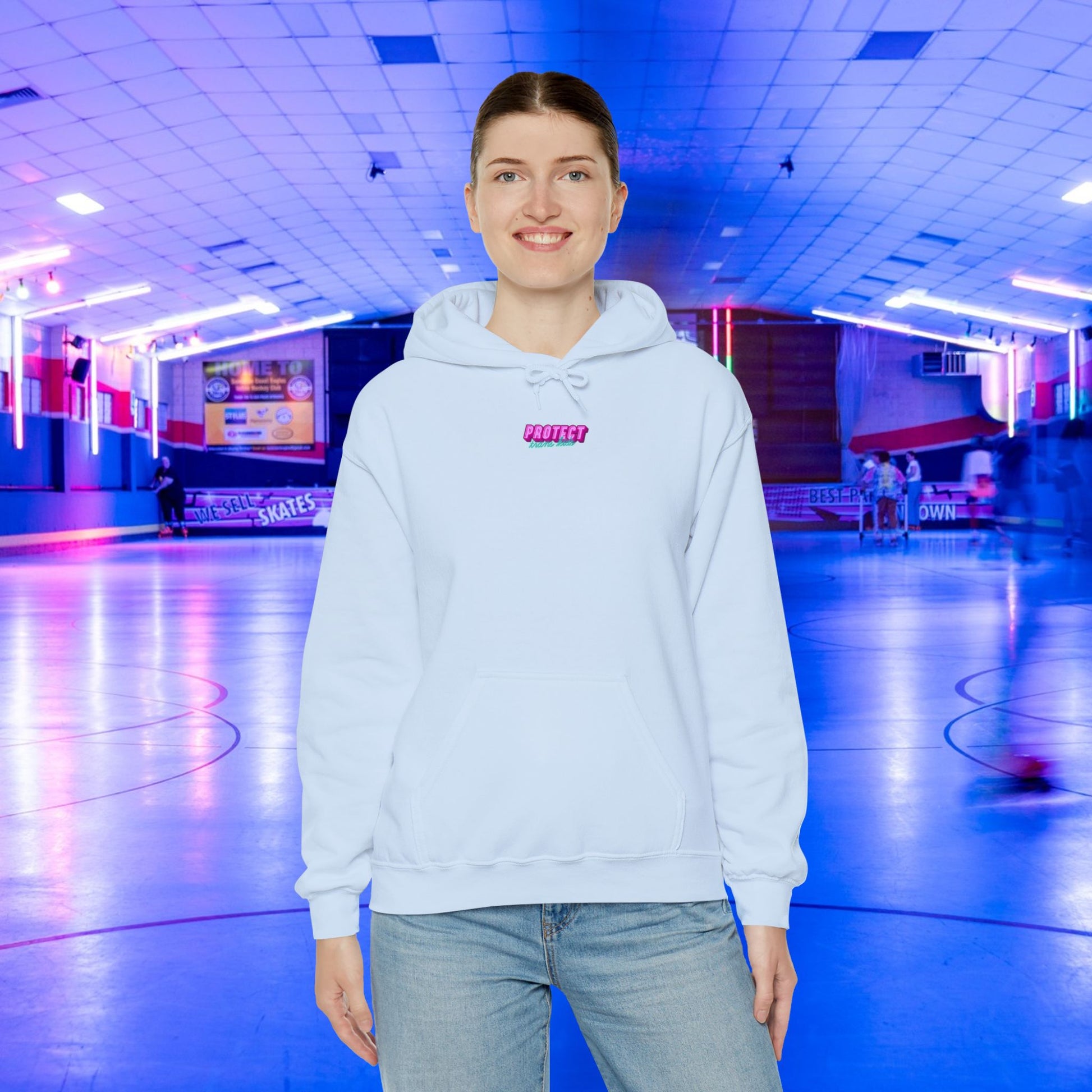 A person in a "Protect Trans Kids" Hoodie stands smiling in an indoor roller skating rink with a bluish-purple glow. Skaters glide under neon lights, creating a lively atmosphere amidst ethically grown cotton hoodie and jeans worn by the individual.