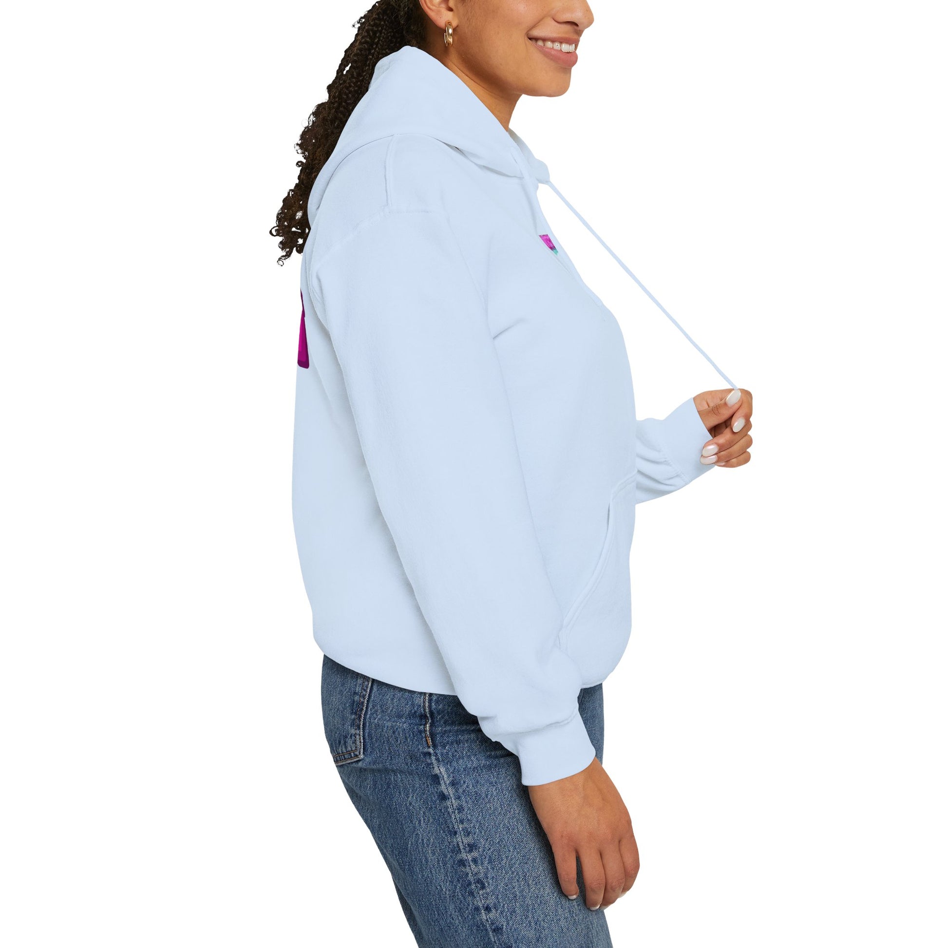 A person in a light blue "Protect Trans Kids" hoodie made from ethically grown cotton and blue jeans smiles while looking to the side. They hold one of the hoodie strings with a pink design partially visible. Their tied-back curly hair adds to their laid-back style.