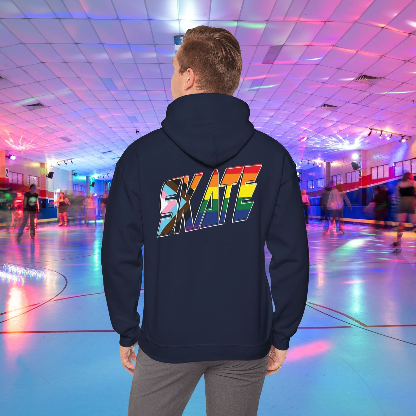 A person with short hair is wearing a GAYSKATE SKATE Progress Pride Hoodie, showcasing a rainbow pride flag gradient "SKATE" design on the back. They are facing away from the camera in a roller skating rink illuminated by vibrant, colorful lights and surrounded by other skaters.