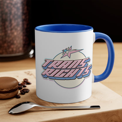 The Trans Rights colorful accent mug by Printify showcases a white coffee mug with a pink handle and vibrant interior, featuring the bold, retro-style text "TRANS RIGHTS" against a circular, off-white background. Above the text is a stylized star with dynamic lines that reflect the colors of the transgender pride flag, creating an energetic sense of movement.