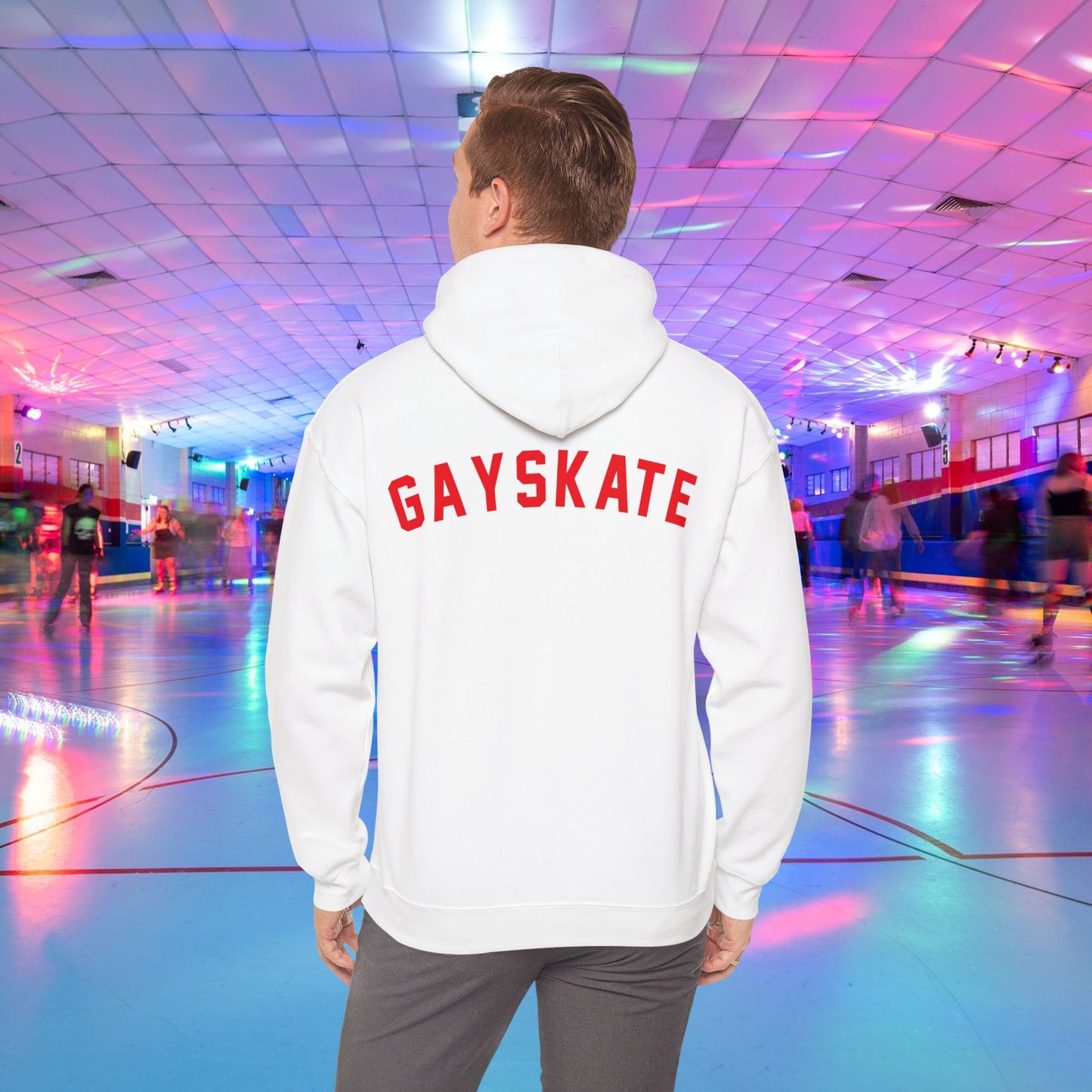 A person wearing a Printify Gay Skate 1982 Retro Hoodie, a high-quality unisex heavy blend hooded sweatshirt, with the word "GAYSKATE" in bold red letters on the back stands in a brightly lit roller skating rink. The rink features colorful lights and skaters in motion, creating a lively atmosphere.