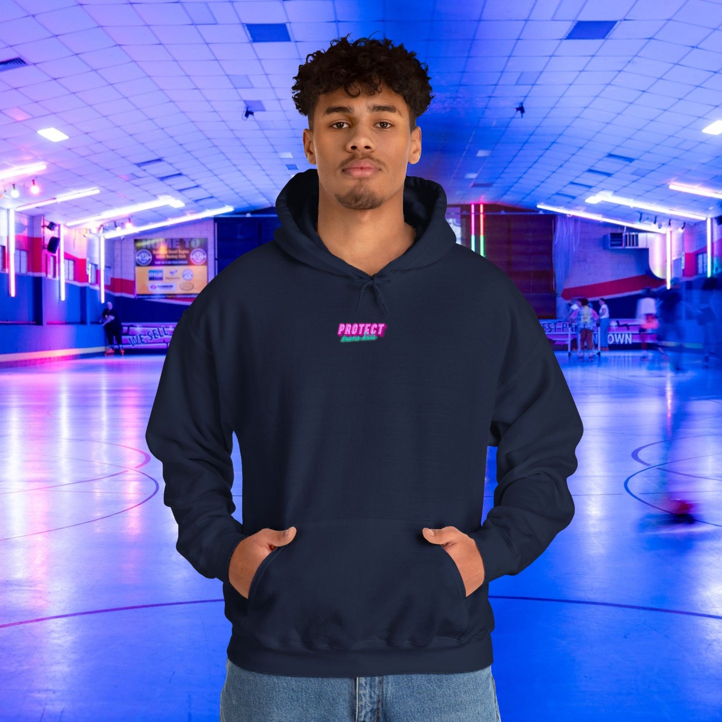 In a dim roller skating rink, a man wears the Protect Trans Kids Hoodie, featuring "PROTECT" in pink on navy. Made from ethically grown cotton, this unisex heavy blend sweatshirt pairs well with the blurred skaters and vibrant lights around him.