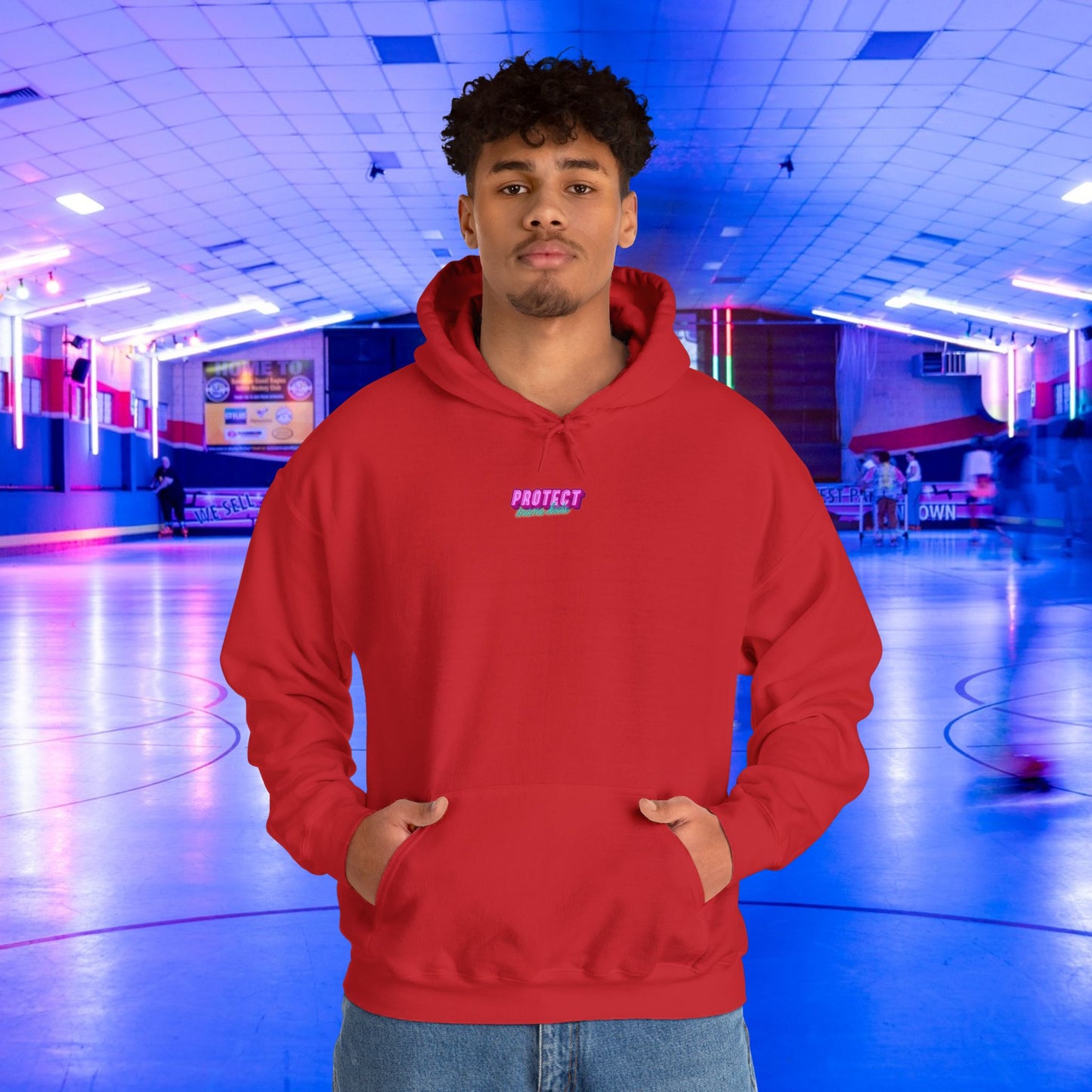 A person wearing the Protect Trans Kids Hoodie in red stands with hands in pockets at an indoor roller skating rink with colorful lights and blurred skaters. This unisex heavy blend sweatshirt, made from ethically grown cotton, features "PROTECT" on the front.