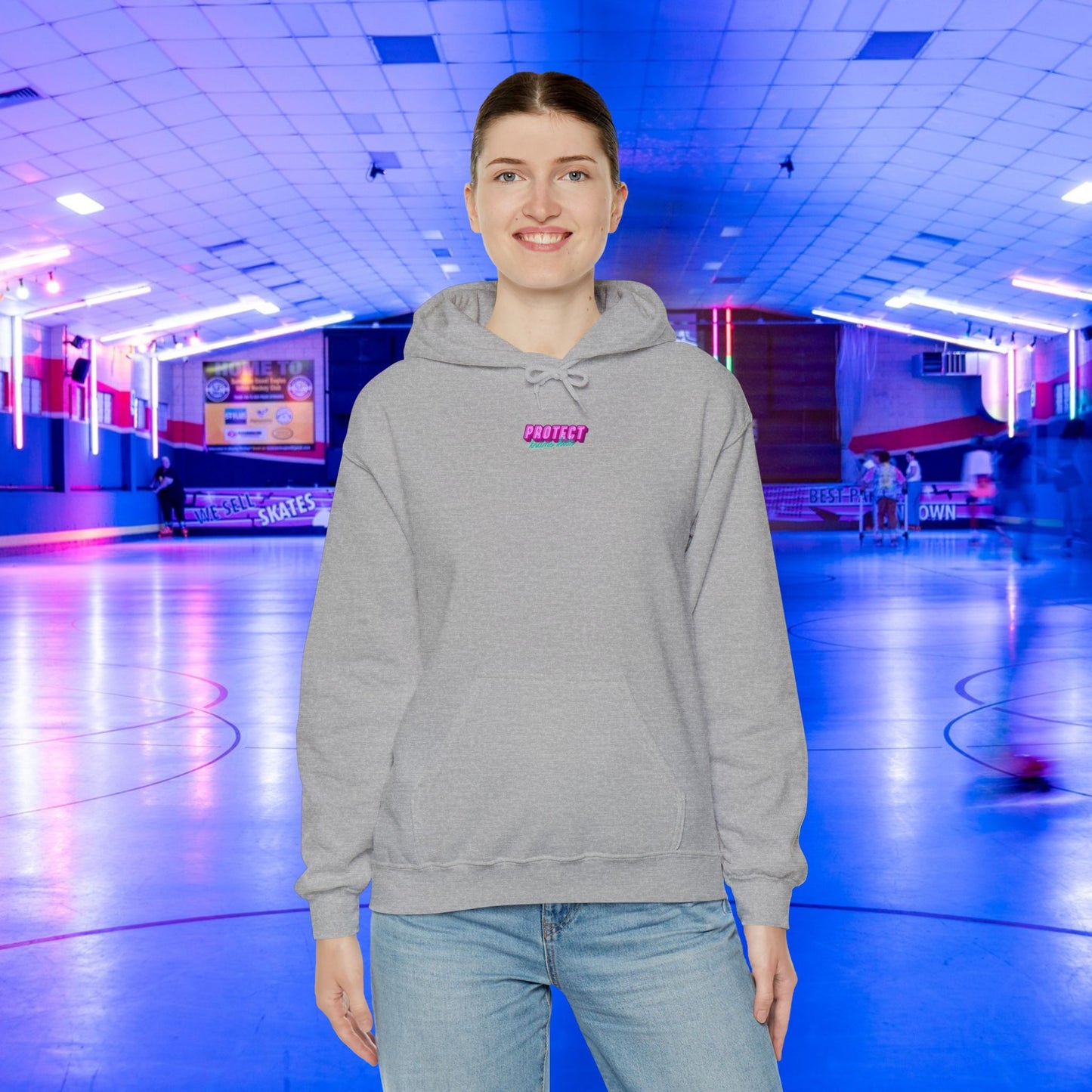 A smiling person wearing the "Protect Trans Kids" hoodie stands in a neon-lit roller skating rink. The gray unisex hoodie, crafted from ethically grown cotton, features "Protect" in pink lettering as blurred skaters move in the background. .