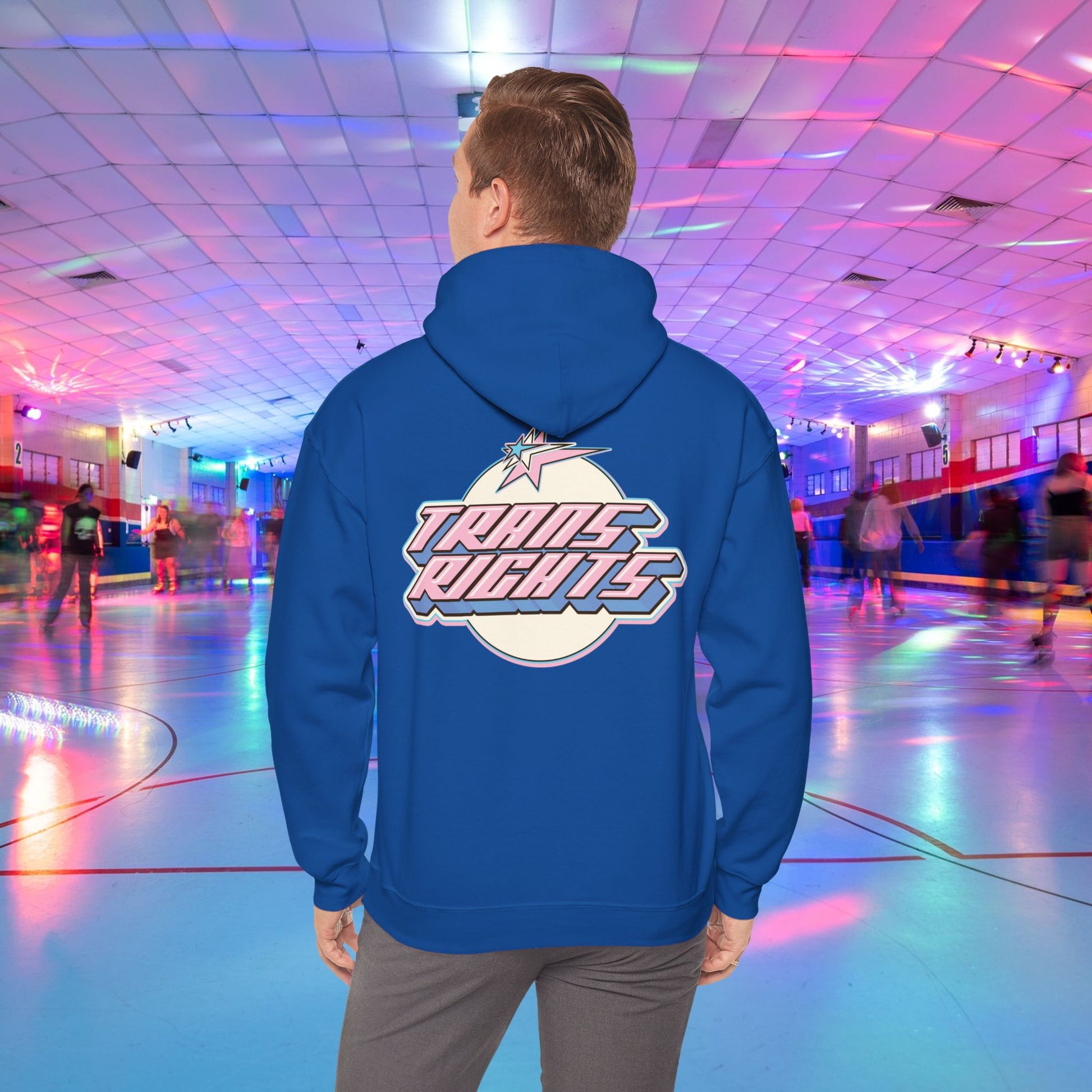 A person wearing a TRANS RIGHTS Hoodie in trans flag colours - Australian Shipping from GAY SKATE stands in a brightly lit roller skating rink, decorated with colorful lights and occupied by people skating in the background.