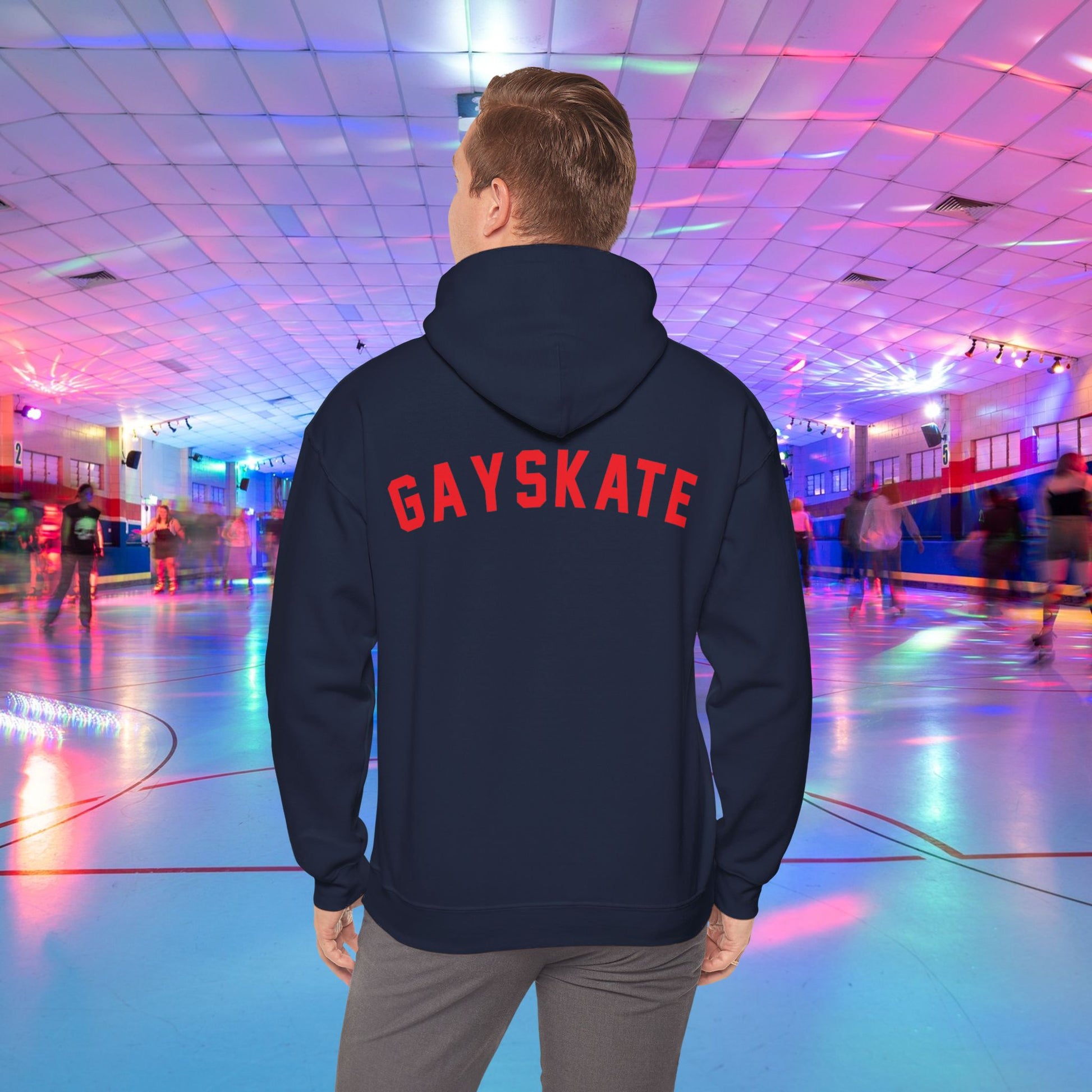 A person wearing a Printify Gay Skate 1982 Retro Hoodie, a high-quality unisex heavy blend hooded sweatshirt, with the word "GAYSKATE" in bold red letters on the back stands in a brightly lit roller skating rink. The rink features colorful lights and skaters in motion, creating a lively atmosphere.