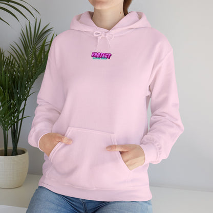 A person wears the Protect Trans Kids Hoodie, a light pink unisex heavy blend sweatshirt with "PROTECT" in purple and black lettering. They stand indoors with hands in the front pocket, surrounded by greenery, highlighting the soft charm of ethically grown cotton.