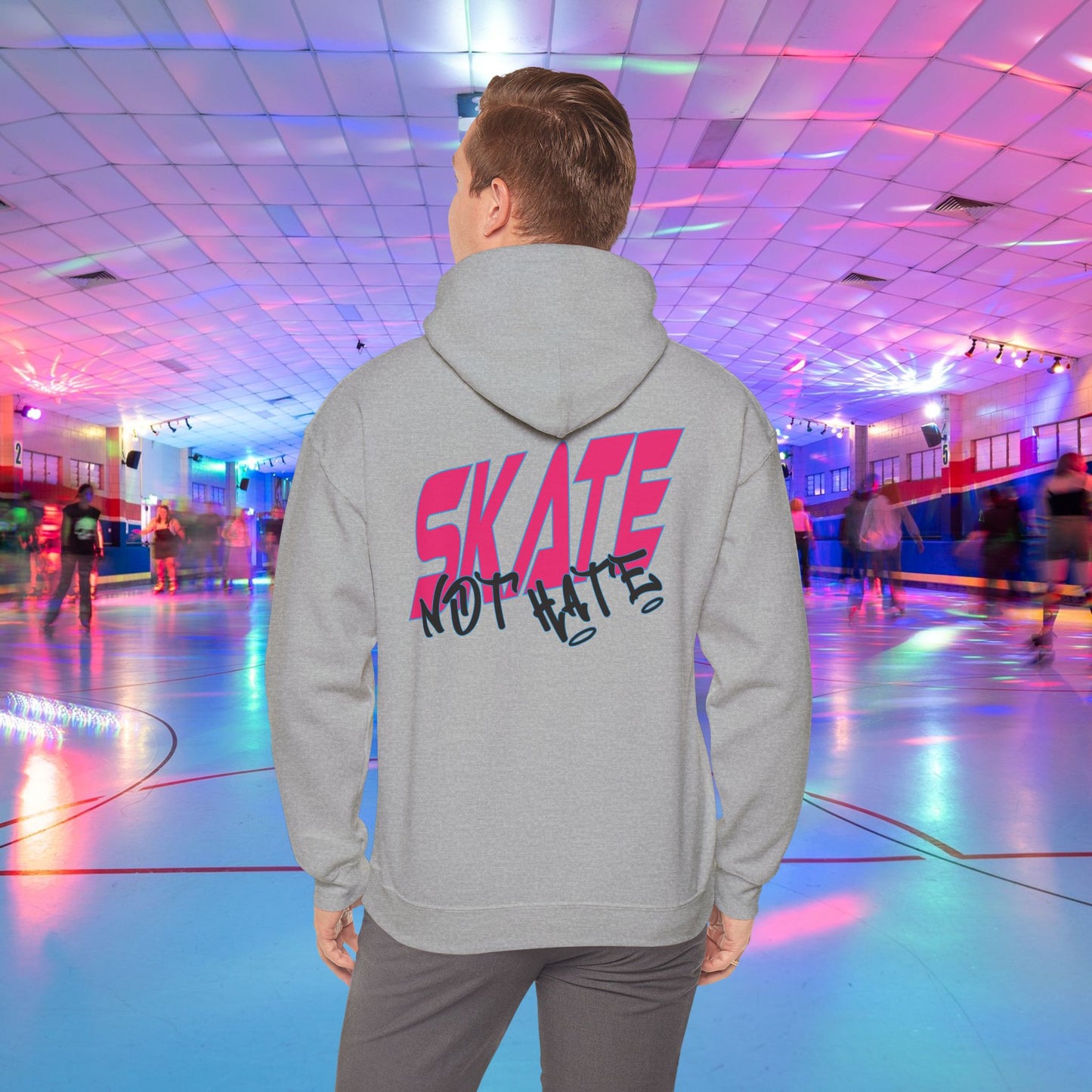 A person wearing a SKATE Not Hate Hoodie from Printify, characterized by its light pink color and high-quality construction, stands in a roller skating rink. The ethically-made unisex heavy blend hoodie, crafted from US-grown cotton, features the words "SKATE NOT HATE" printed on the back. The rink is adorned with colorful lights and has a shiny floor, with other skaters gliding in the background.