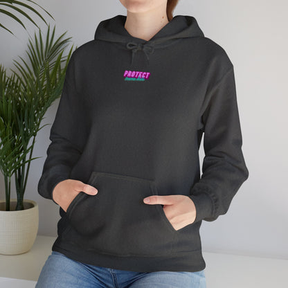 Wearing the "Protect Trans Kids Hoodie," made from ethically grown cotton with "PROTECT" in pink on the front, a person rests their hands in the front pocket, standing near a plant against a light background.