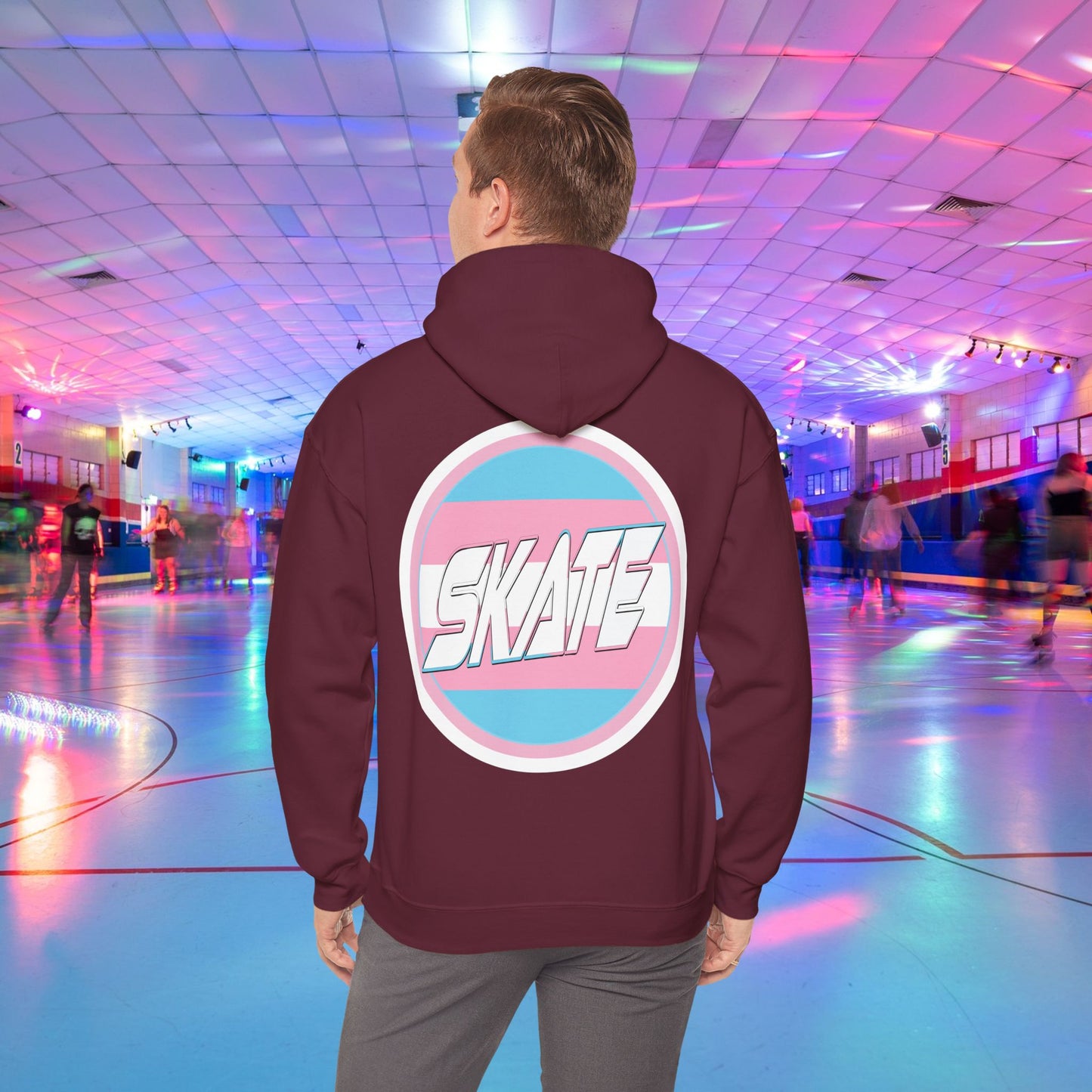 A person wearing a high-quality, unisex "Gay Skate Original - Trans Edition Hoodie" from Printify, featuring a large "SKATE" logo on the back, stands in a colorful roller skating rink. The rink is illuminated with multicolored lights and has several people skating in the background. The overall atmosphere is lively and vibrant.