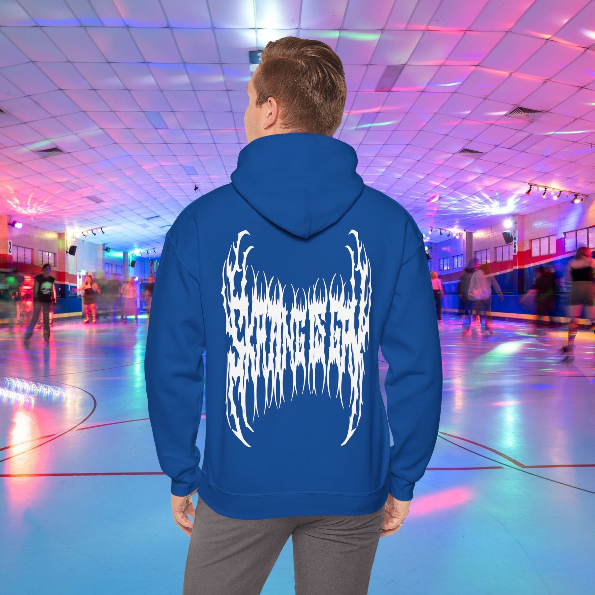 A person wearing the "SKATING IS GAY but make it death metal" hoodie stands in a brightly lit roller skating rink. The rink is adorned with colorful lights, and other people are skating in the background.