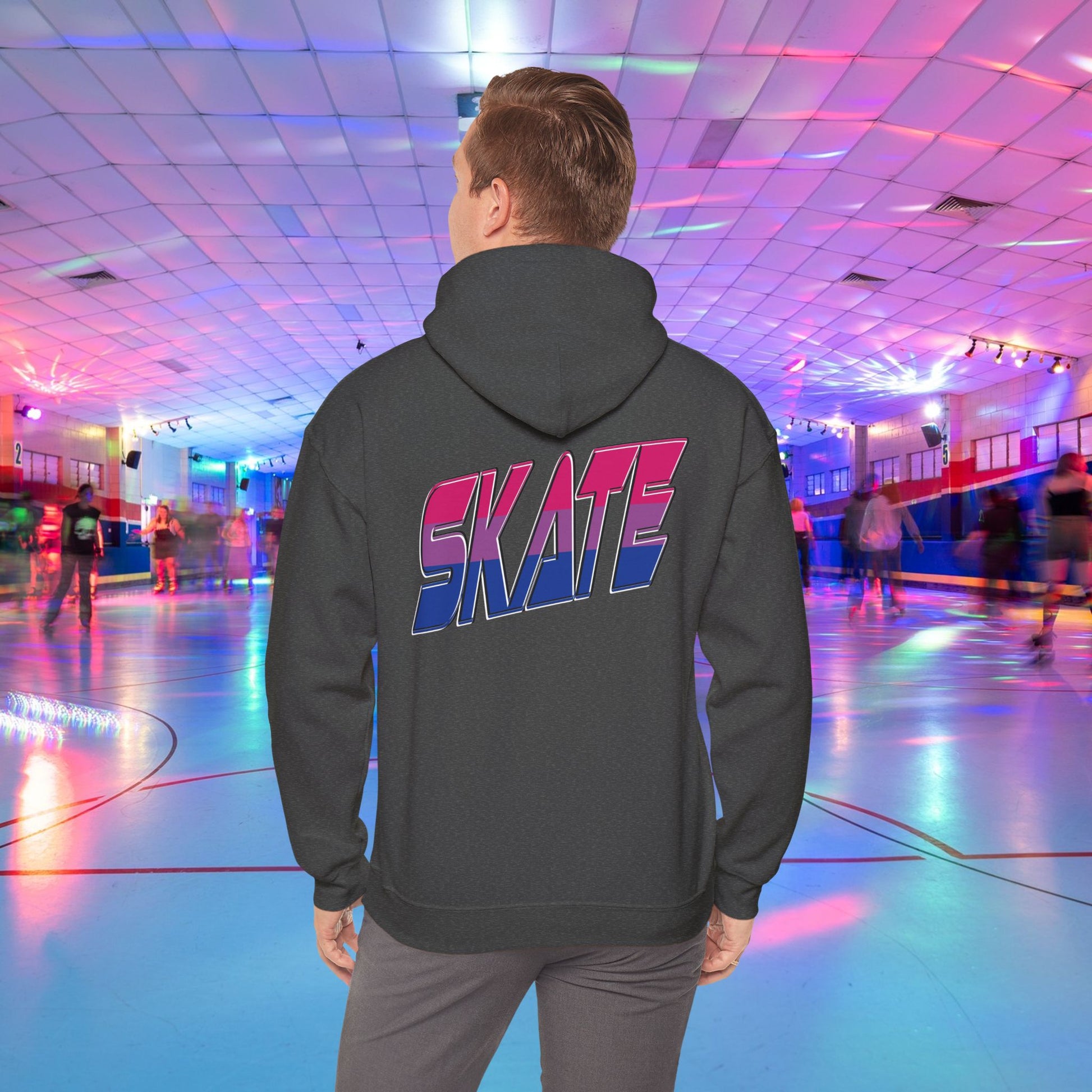 A person in a SKATE Bisexual Pride Hoodie from Printify, with bold and colorful "SKATE" lettering on the back, stands in a brightly lit roller skating rink. The background features skaters gliding on a shiny floor under a ceiling with vibrant, multicolored lights. This unisex heavy blend hooded sweatshirt showcases original designs and is available for Australian shipping.