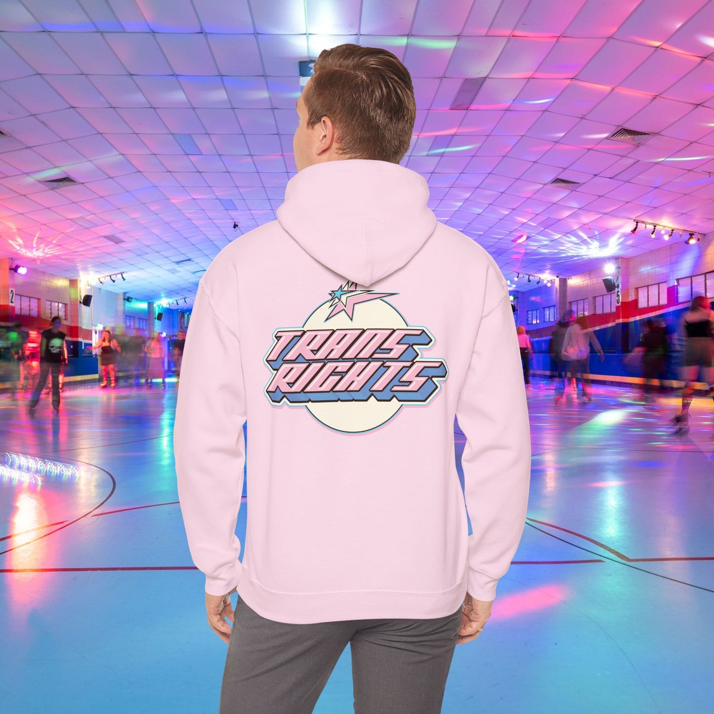 A person wearing a TRANS RIGHTS Hoodie in trans flag colours - Australian Shipping from GAY SKATE stands in a brightly lit roller skating rink, decorated with colorful lights and occupied by people skating in the background.