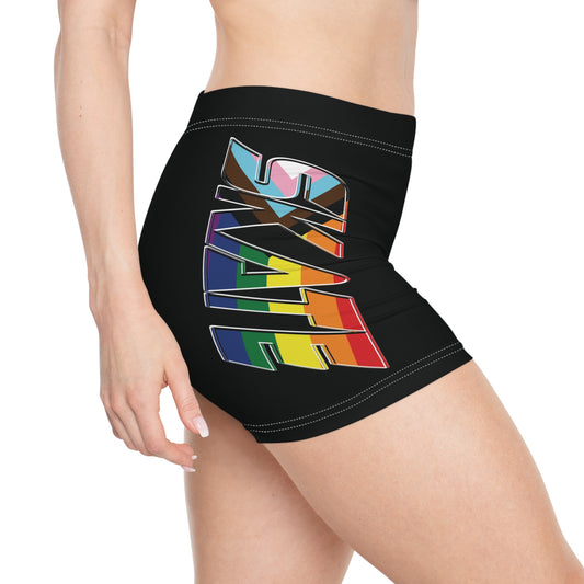 A person is seen from the side wearing the Skate Pride Flag Short Shorts by gay skate, which are black and have "SKATE" printed in large, colorful letters on the side in the rainbow pride flag. These shorts, made from a polyester spandex blend, feature a wide waistband and offer a snug fit for comfort and durability. The background is plain white.