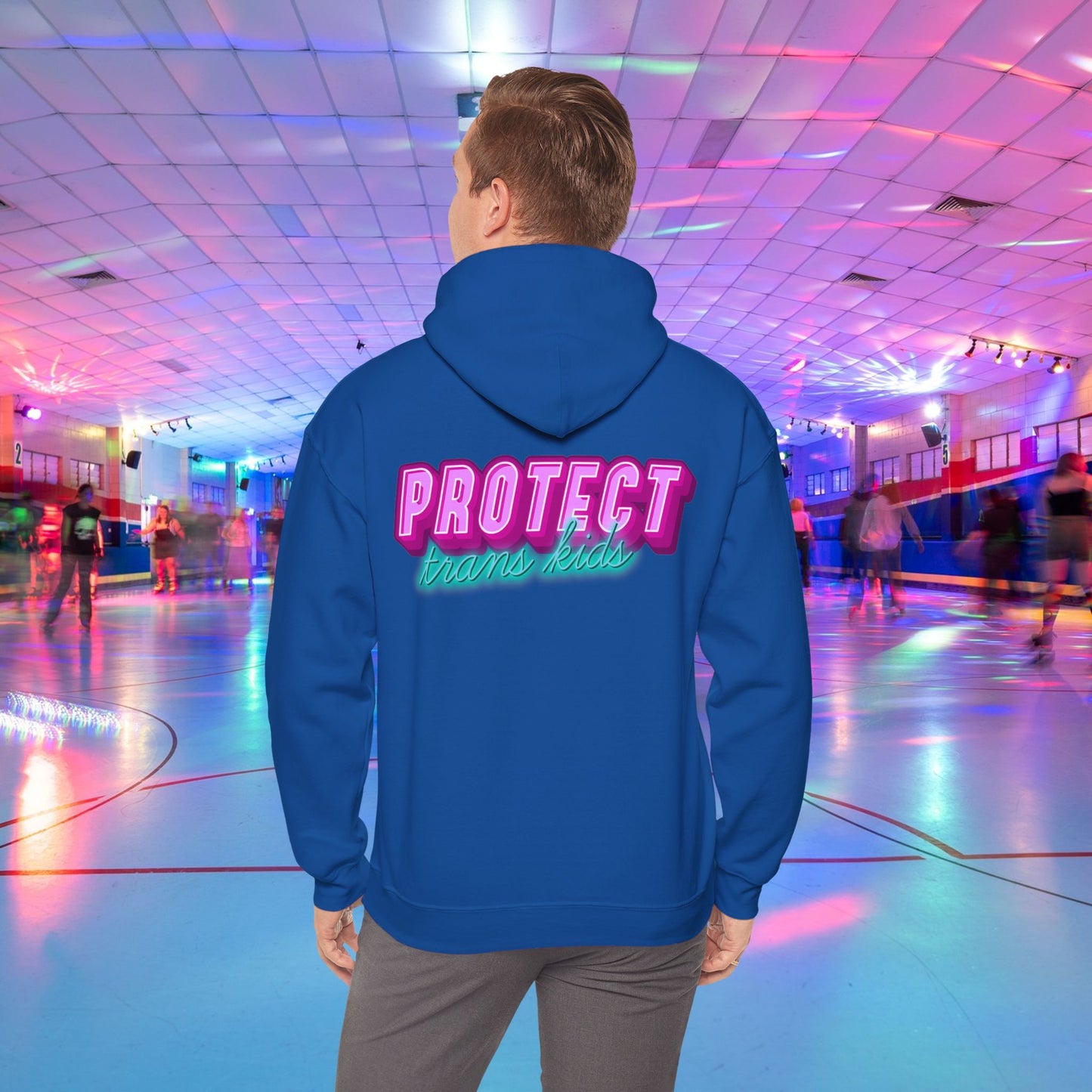 A person is at a roller skating rink, sporting a high-quality Protect Trans Kids Hoodie from Printify, crafted from ethically grown cotton, with "PROTECT trans kids" boldly and colorfully emblazoned on the back. The rink is bathed in vibrant, colorful lights, and other skaters can be seen gliding in the background.