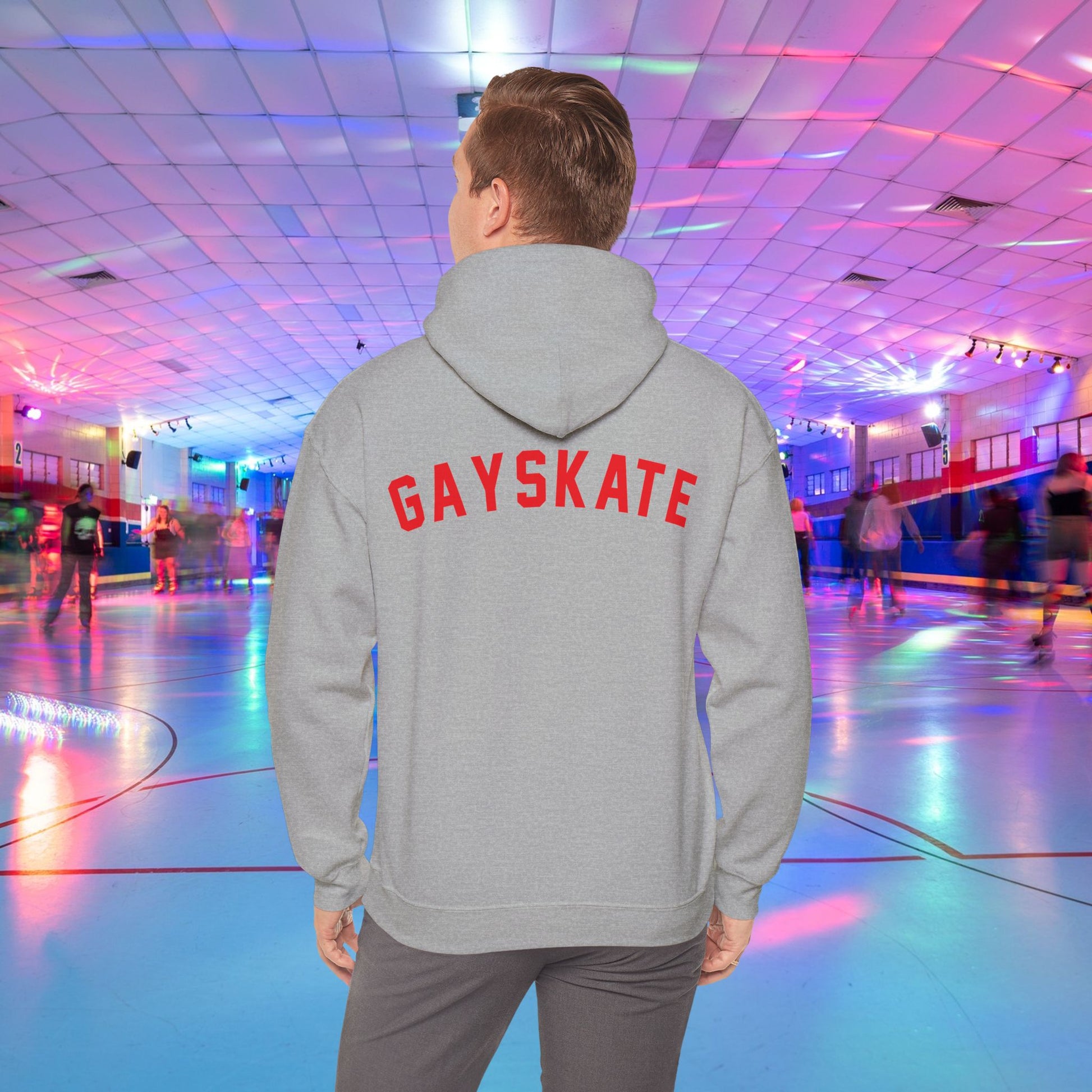 A person wearing a Printify Gay Skate 1982 Retro Hoodie, a high-quality unisex heavy blend hooded sweatshirt, with the word "GAYSKATE" in bold red letters on the back stands in a brightly lit roller skating rink. The rink features colorful lights and skaters in motion, creating a lively atmosphere.