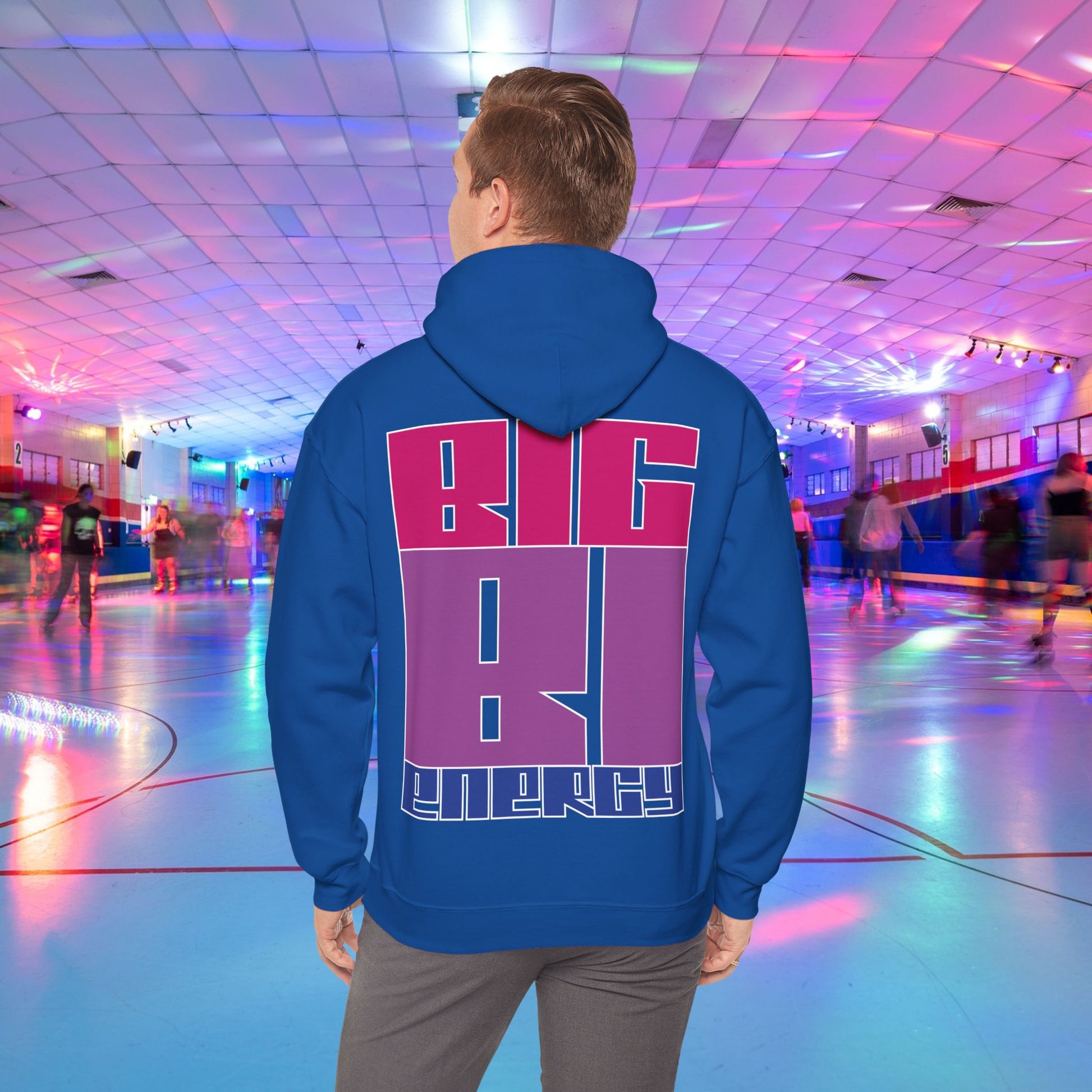 A person with light brown hair is wearing a high-quality BIG BI ENERGY Hoodie from GAY SKATE, featuring bold, Bisexual Pride Flag colour letters on the back. They are standing in a roller skating rink illuminated with vibrant, colorful lights. The cozy feel and warmth of this trans and queer-owned brand is evident.