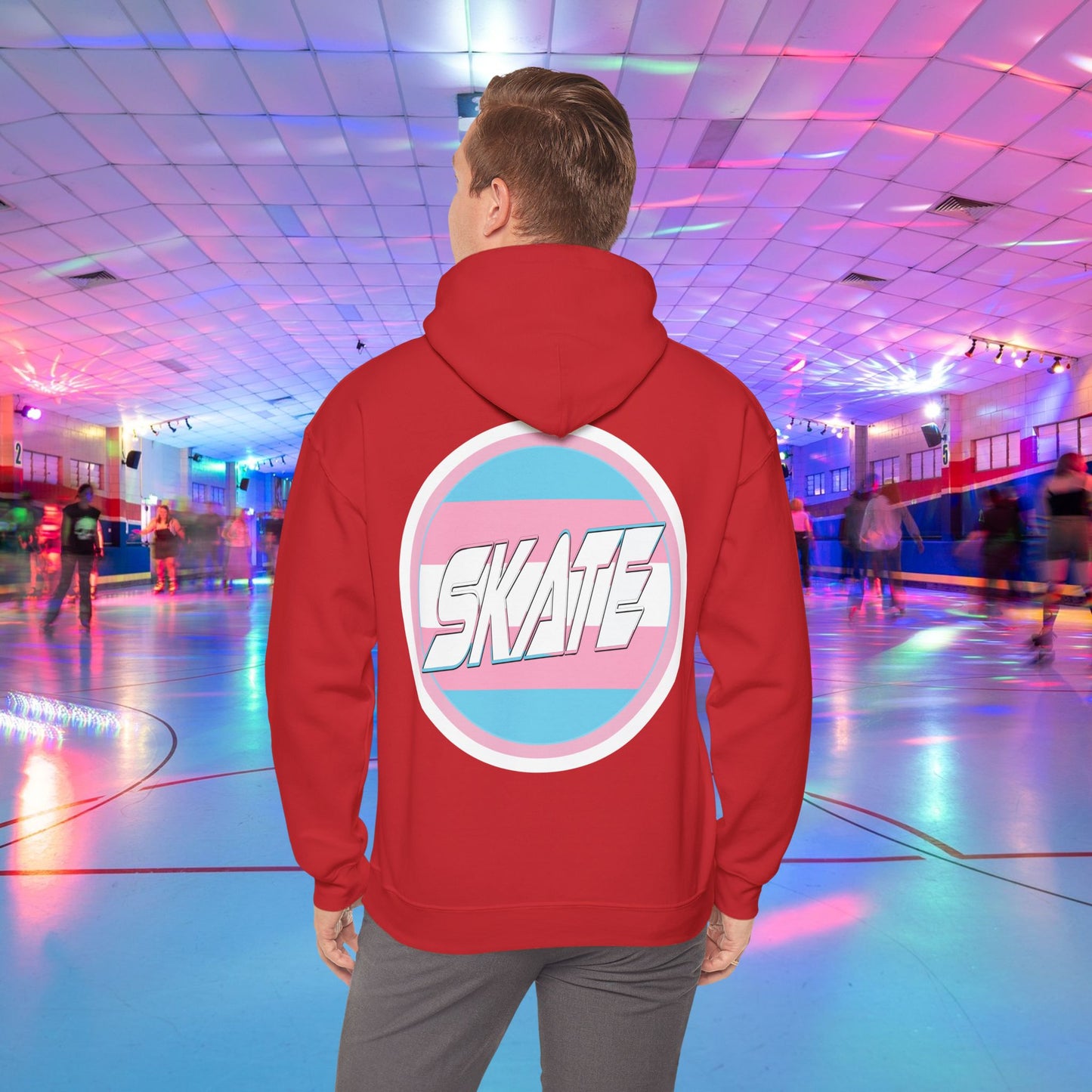 A person wearing a Printify SKATE Trans Flag round logo Hoodie, featuring the word "SKATE" inside a blue and pink circular graphic, stands at the center of a colorful roller skating rink with vibrant lights and other skaters in the background.